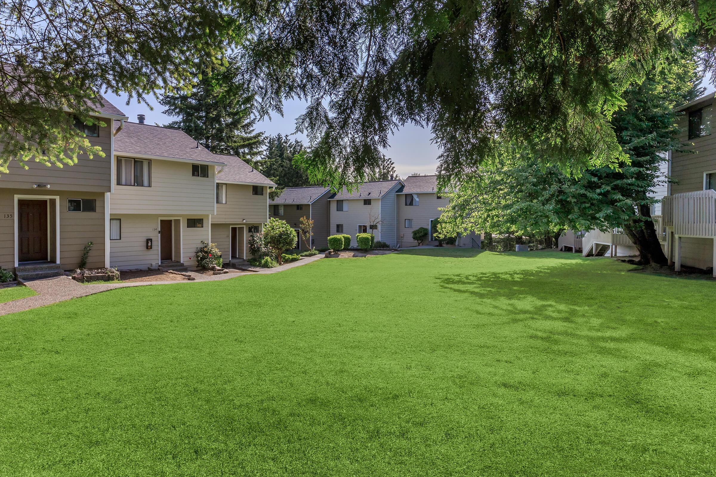 A grassy courtyard surrounded by residential buildings, with a few trees providing shade. The scene features well-maintained lawns and pathways leading to the entrances of the homes, suggesting a peaceful neighborhood setting. The sky is clear and bright, indicating a sunny day.