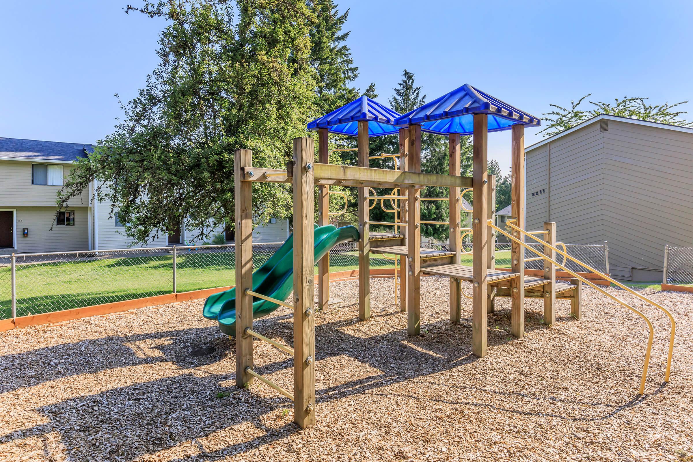 A colorful playground featuring a wooden structure with a green slide, blue canopies, and climbing areas. The playground is situated on a bed of wood chips, surrounded by a fenced area and residential buildings in the background, under a clear blue sky.