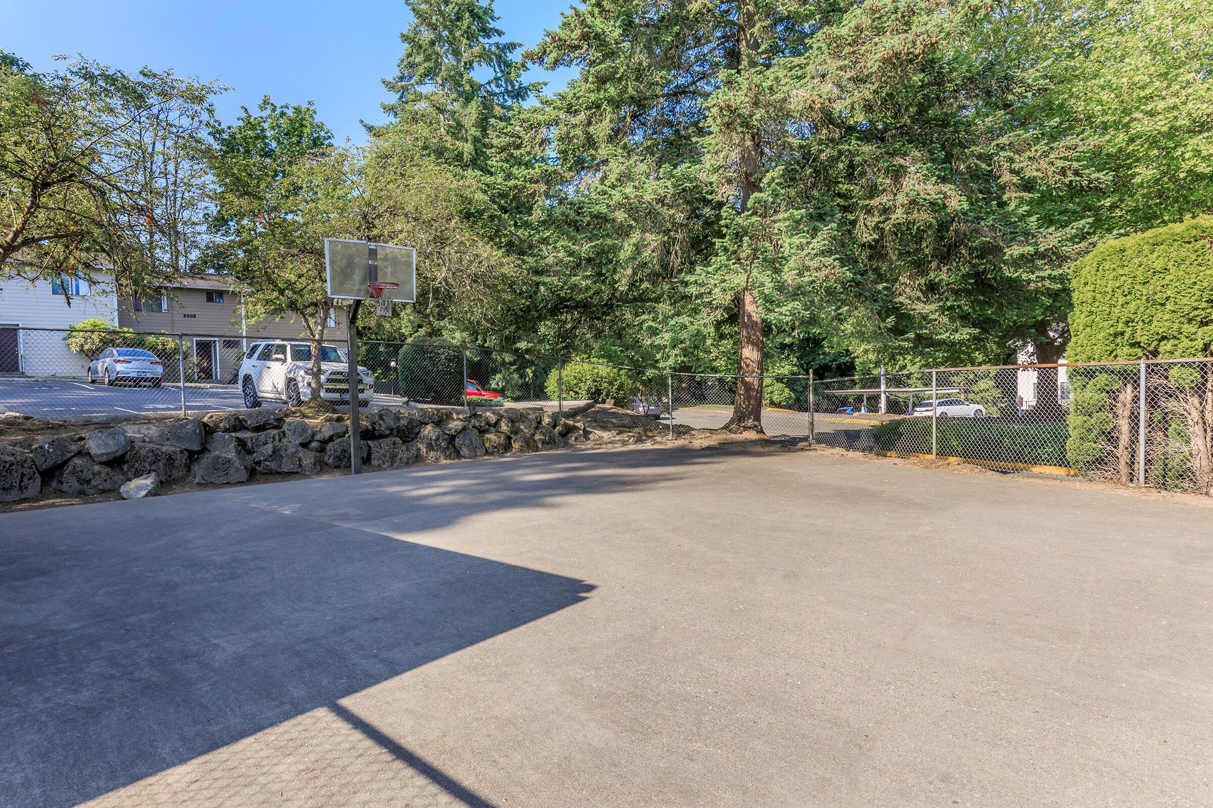A quiet outdoor basketball court surrounded by trees, with a chain-link fence and a stone wall along one side. A parked car is visible in the background, indicating a residential area nearby.
