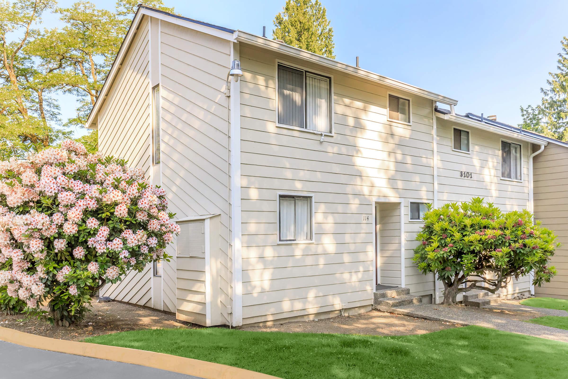 A two-story residential building with light-colored siding, featuring a pathway, windows, and two front doors. There are landscaped bushes and vibrant flowering plants in front of the building, with trees in the background indicating a sunny day.