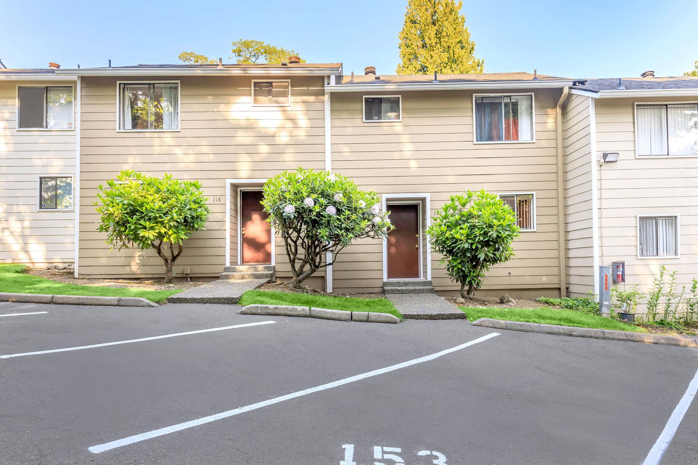 Two-story apartment building with beige siding, featuring two front doors flanked by blooming bushes. The parking area in front has marked parking spots, and a well-maintained lawn is visible. The scene is set in a sunny outdoor environment with clear blue skies.