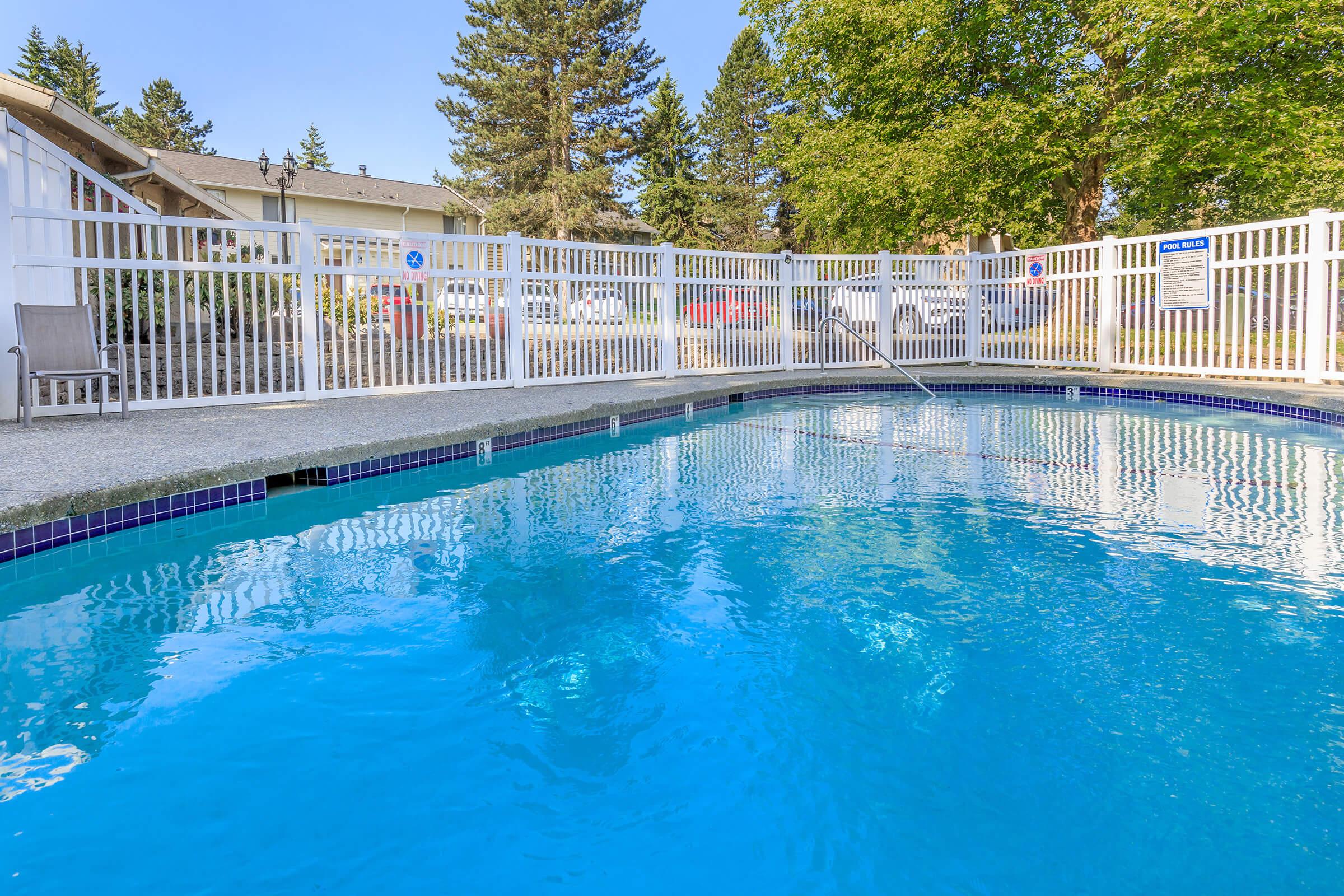 A sparkling blue swimming pool enclosed by a white fence, with a clear sky and trees in the background. Sunlight reflects off the water's surface, creating a serene atmosphere. A few lounge chairs are positioned nearby, adding to the inviting space for relaxation.