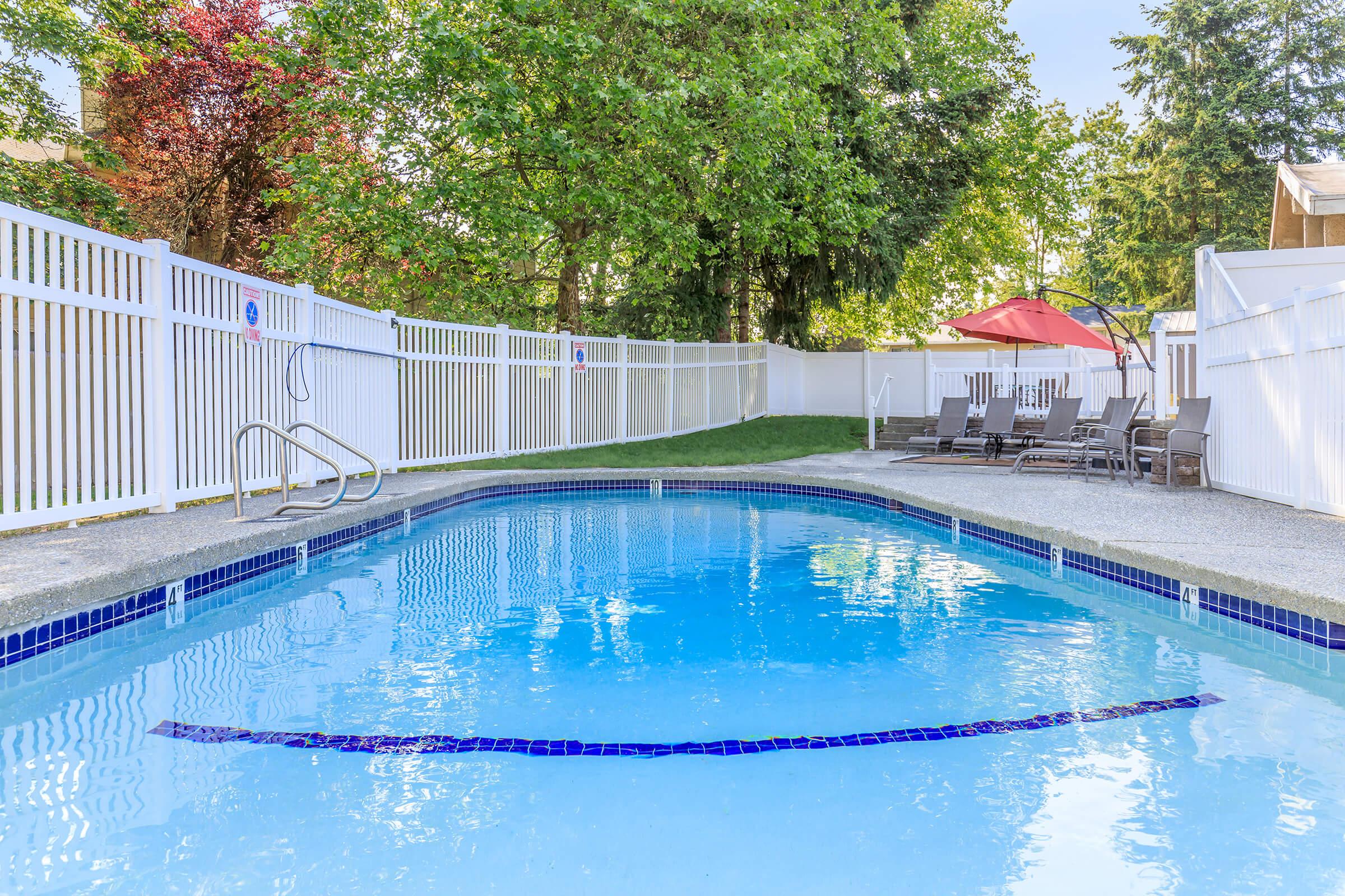 A clear swimming pool with a blue tiled border, surrounded by a white picket fence. Lounge chairs line one side of the pool, with an umbrella providing shade nearby. Lush green trees provide a backdrop, and the area appears well-maintained and inviting for relaxation.