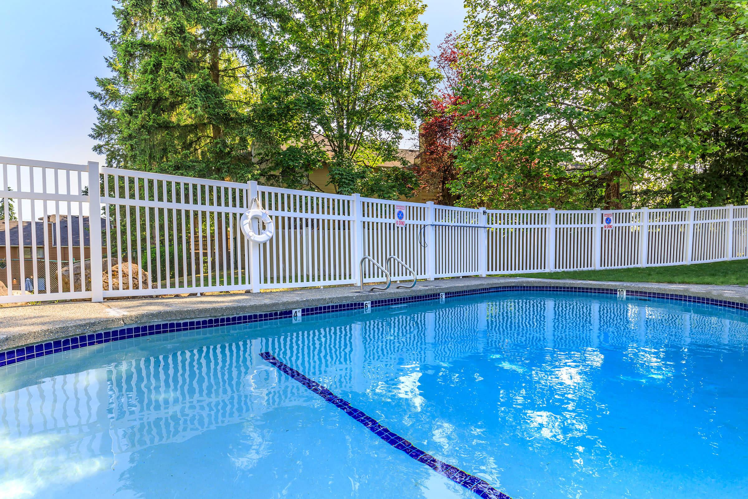 A clear swimming pool surrounded by a white fence, with trees and greenery in the background. The water is calm and reflects the sky. A lifebuoy is attached to the fence, and the pool has stairs leading in. The setting is serene, ideal for relaxation.