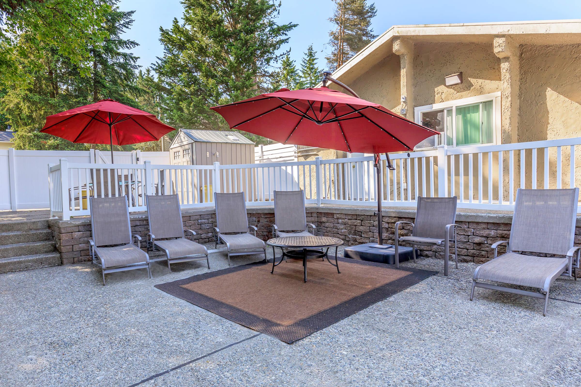 A cozy outdoor seating area featuring six lounge chairs under two large red umbrellas, with a small round table at the center. The setting is complemented by a stone wall and a light-colored house in the background, surrounded by tall trees. Perfect for relaxation in a tranquil environment.