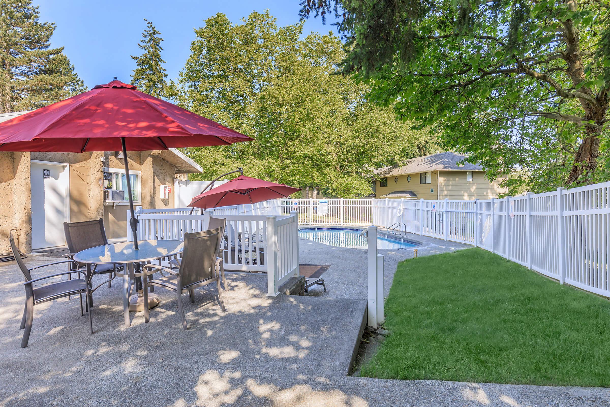 A backyard pool area featuring a swimming pool surrounded by a safety fence. There are two red umbrellas providing shade over a circular table with chairs. Lush green trees are visible in the background, adding to the outdoor ambiance. A manicured lawn is nearby, creating a relaxing atmosphere.