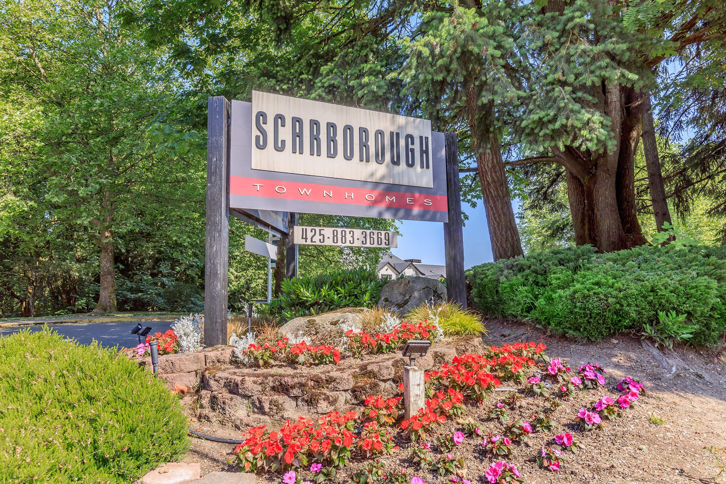 Sign for Scarborough Townhomes, featuring the name prominently displayed at the top, with a phone number below. The sign is surrounded by lush greenery and colorful flower beds, creating an inviting entrance to the residential community.
