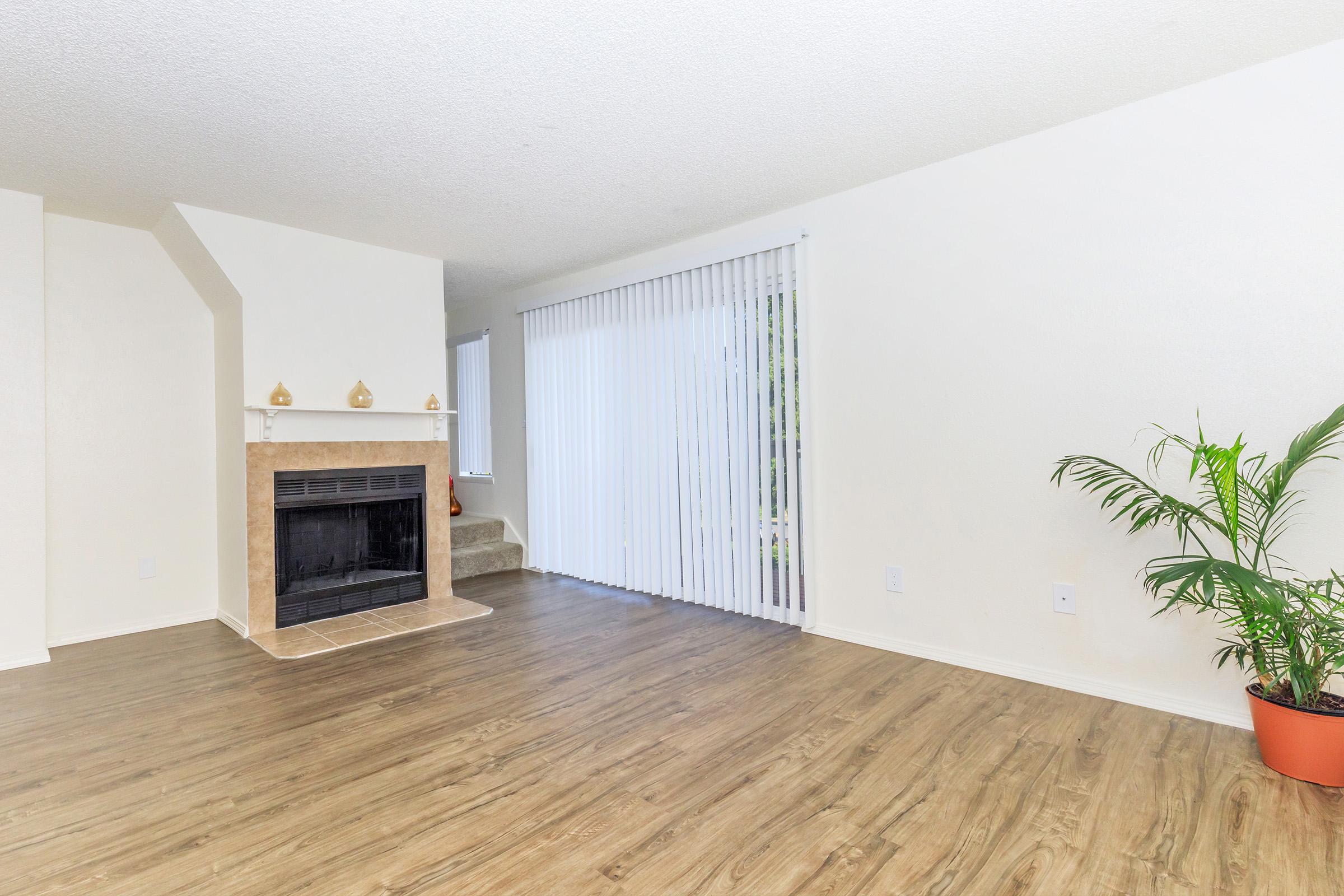 Spacious living room with a tiled fireplace, light hardwood flooring, white walls, and vertical blinds. A potted plant adds a touch of greenery. Natural light flows in from the large window, creating a bright and inviting atmosphere.