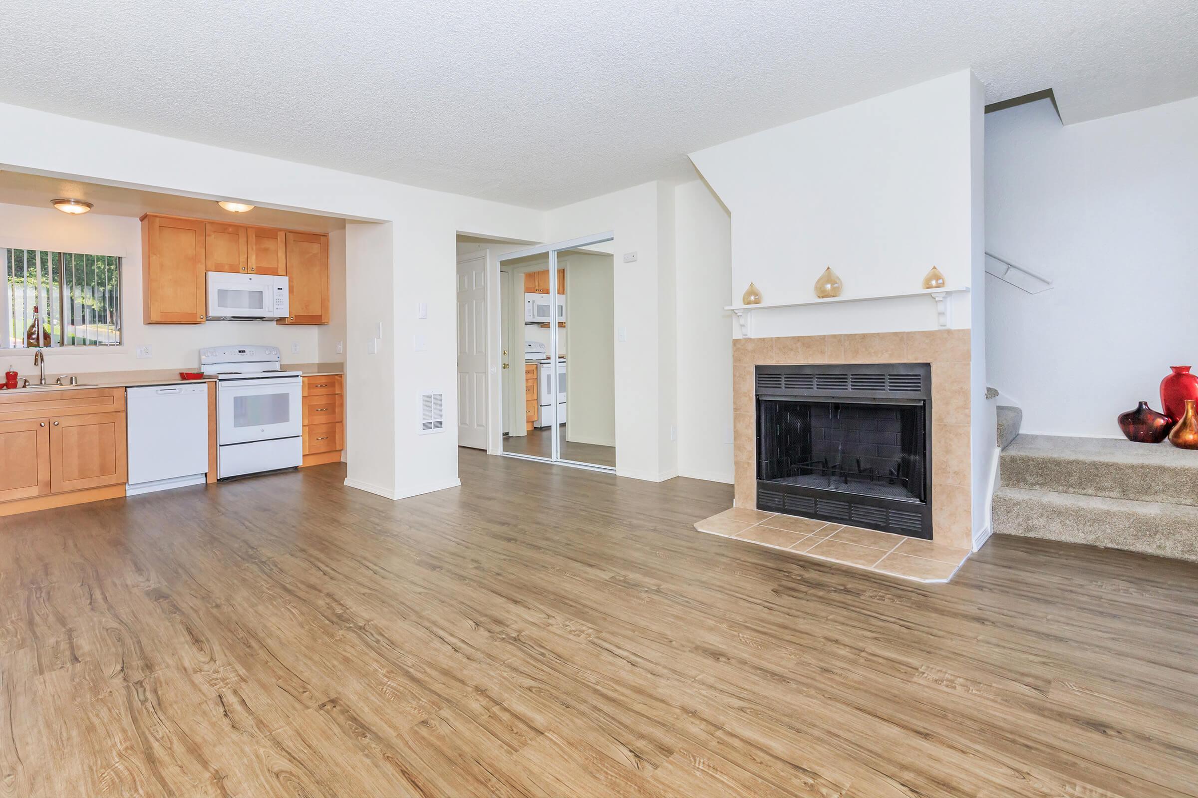 A spacious living area featuring a modern kitchen with wooden cabinets and white appliances. A cozy fireplace is located in the living room, complemented by a carpeted staircase. Large windows provide natural light, enhancing the warm and inviting atmosphere of the space.