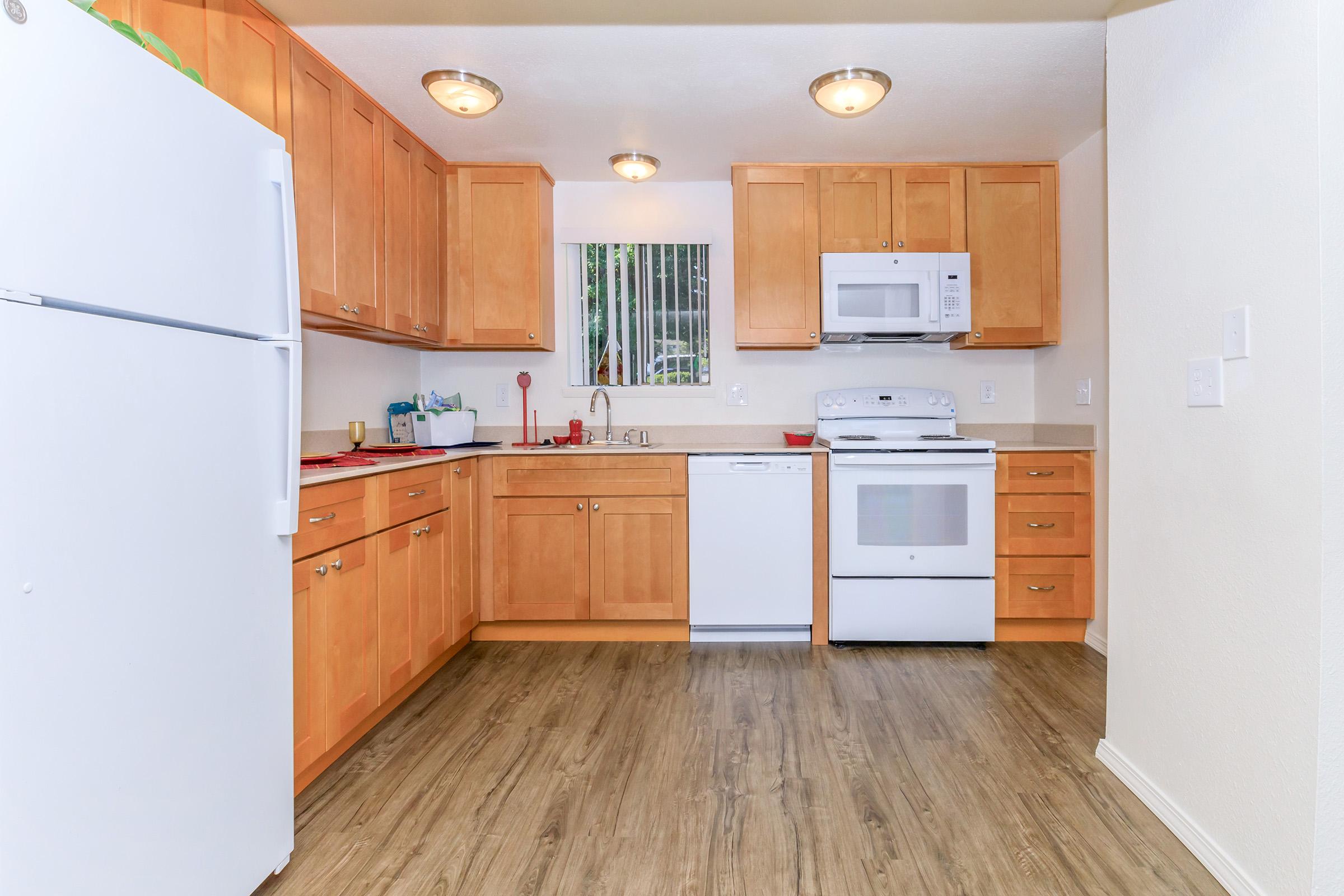 A modern kitchen featuring light wood cabinets, white appliances including a refrigerator, microwave, and dishwasher. The countertop is equipped with utensils and colorful items. The flooring is a warm wood-like material, and there is a window with natural light coming in.