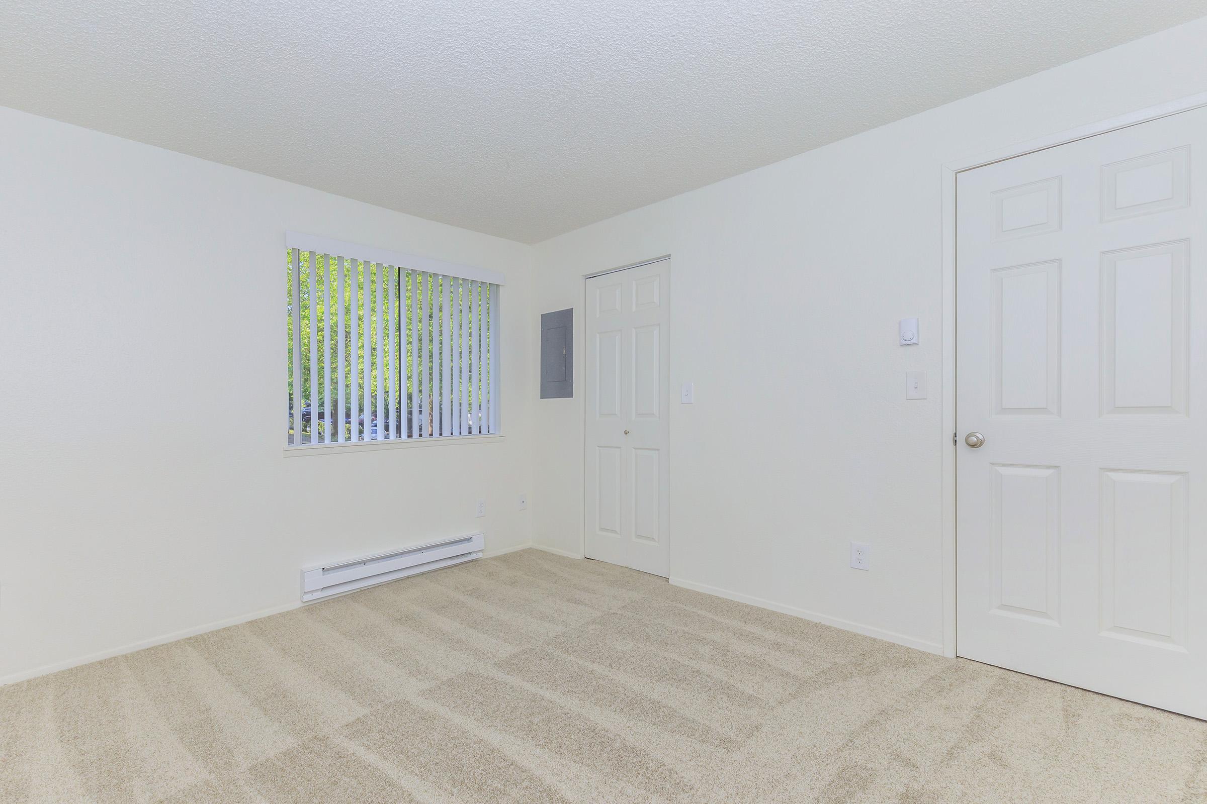 A spacious, empty room with light-colored walls and plush beige carpeting. There is a window with vertical blinds, allowing natural light to enter. A door is visible on the right side of the room, and a small heating unit is mounted along the wall beneath the window. The overall atmosphere is bright and inviting.
