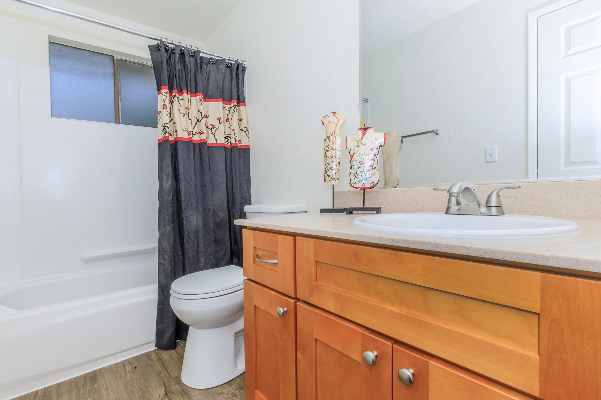 A bright and clean bathroom featuring a wooden vanity with a sink, a toilet, and a shower area. The shower curtain is dark gray with a floral design at the top. The walls are light-colored, and there is a window providing natural light. Overall, the space is modern and well-maintained.