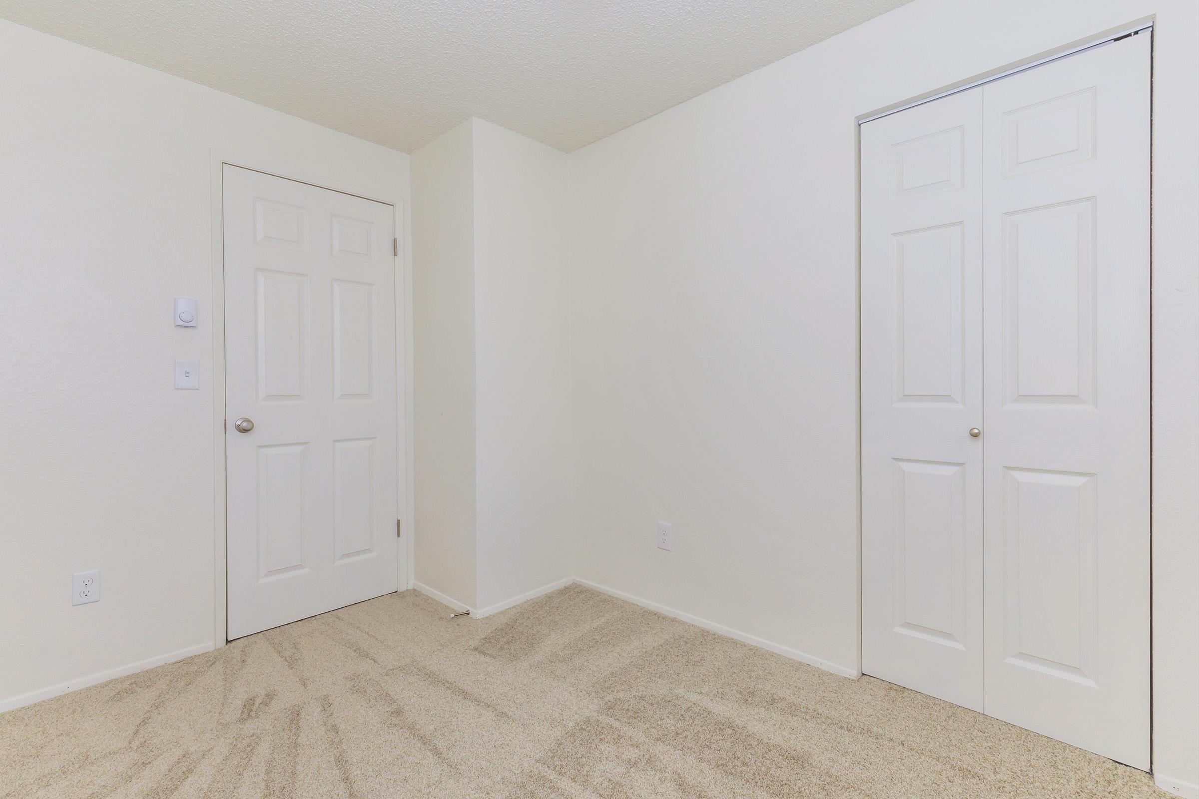 A simple, empty room featuring light beige carpet and plain white walls. Two closed doors are visible on the left and right sides, both with a standard panel design. The room is well-lit, creating a bright and neutral atmosphere, suitable for various uses.