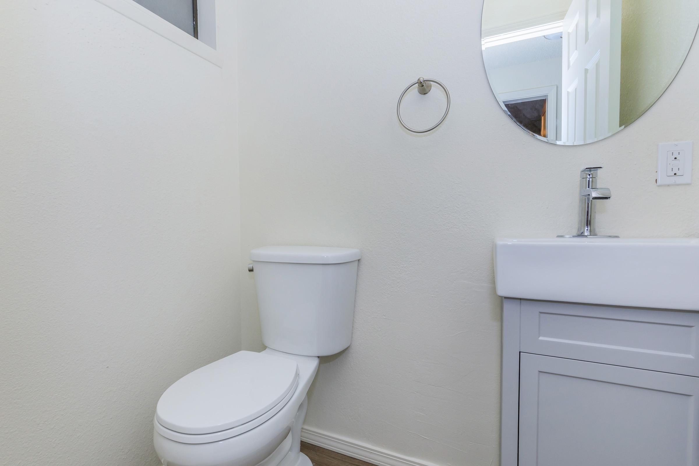 A small bathroom featuring a white toilet, a round mirror above a sink with a modern faucet, and light-colored walls. A cabinet below the sink adds storage space, and the room has a simple, clean design with wood flooring.