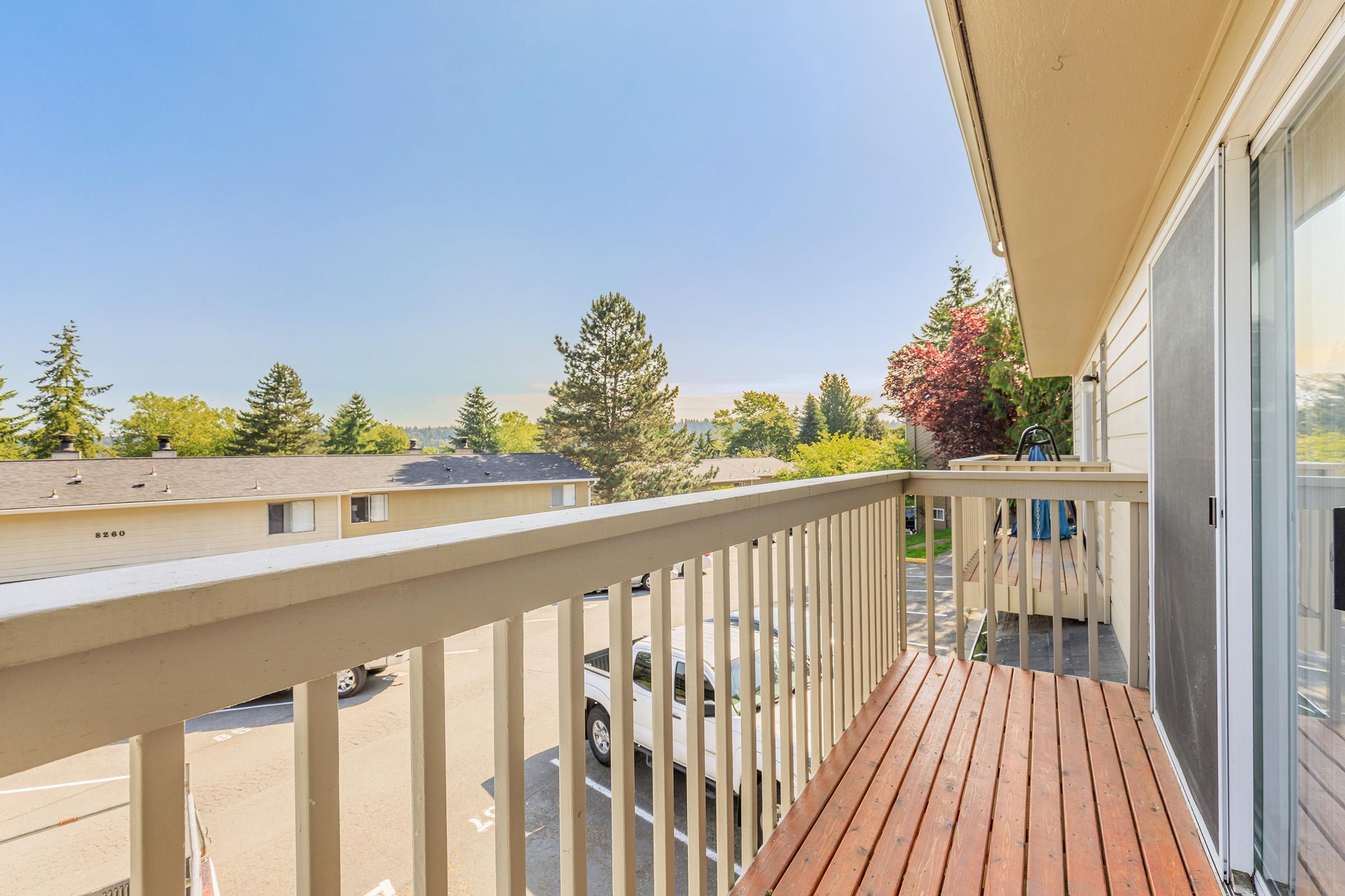 View from a balcony overlooking a residential area. The scene features trees and buildings under a clear blue sky. The balcony has a wooden railing and floor, with a glimpse of parked cars below. The sunlight creates a warm, inviting atmosphere.