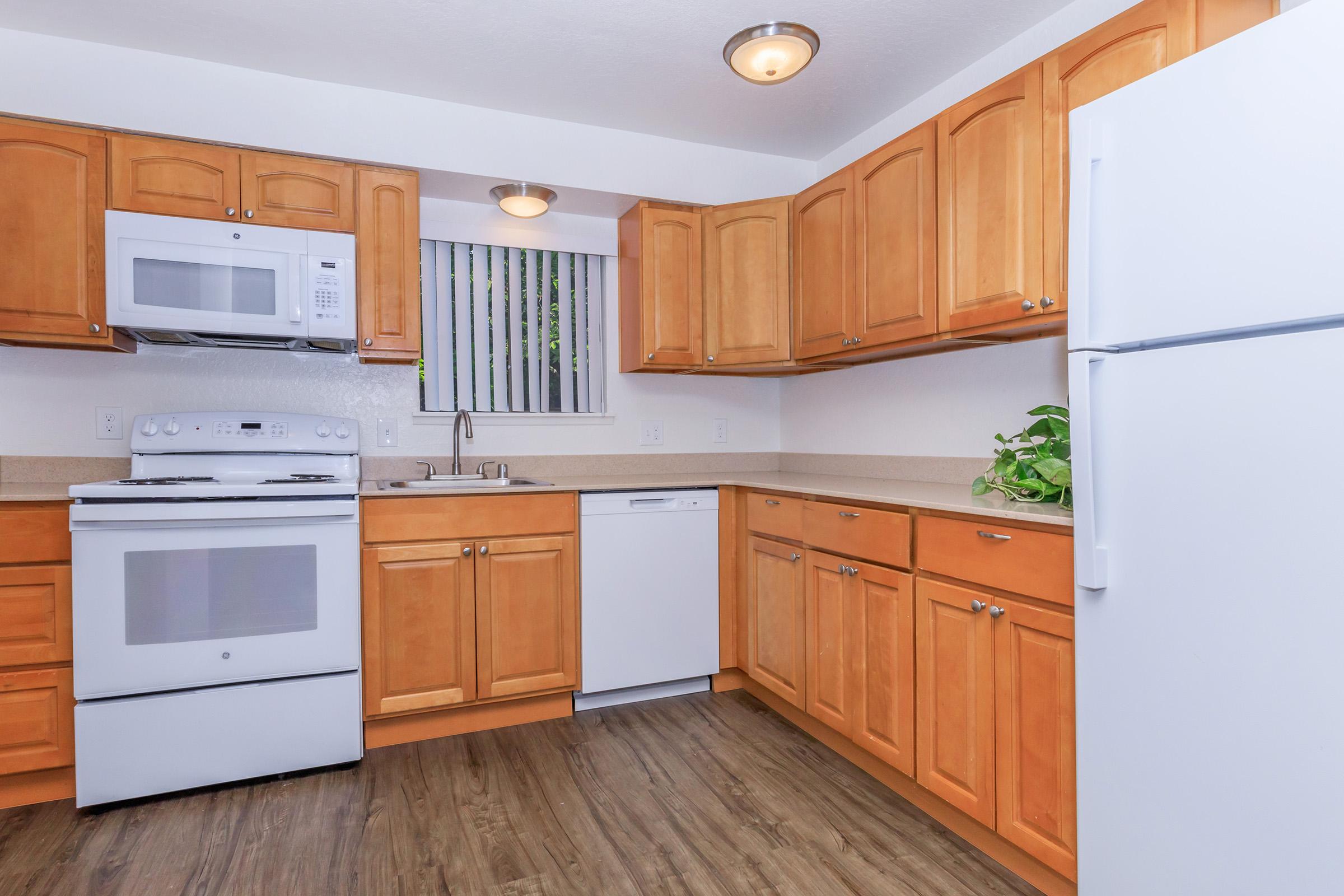 A modern kitchen featuring wooden cabinets, a white stove and microwave, a dishwasher, and a refrigerator. The countertops are light-colored, and there's a window with blinds. The flooring is a dark wood-like laminate, providing a warm and inviting atmosphere. A small plant adds a touch of greenery.