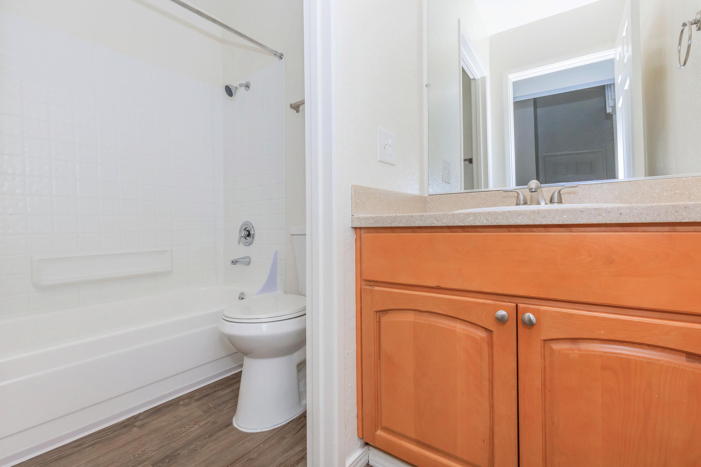 A clean, bright bathroom featuring a bathtub with a shower, a white toilet, and a vanity with wooden cabinetry. Natural light is coming in from a nearby doorway, enhancing the spacious and fresh atmosphere of the space.