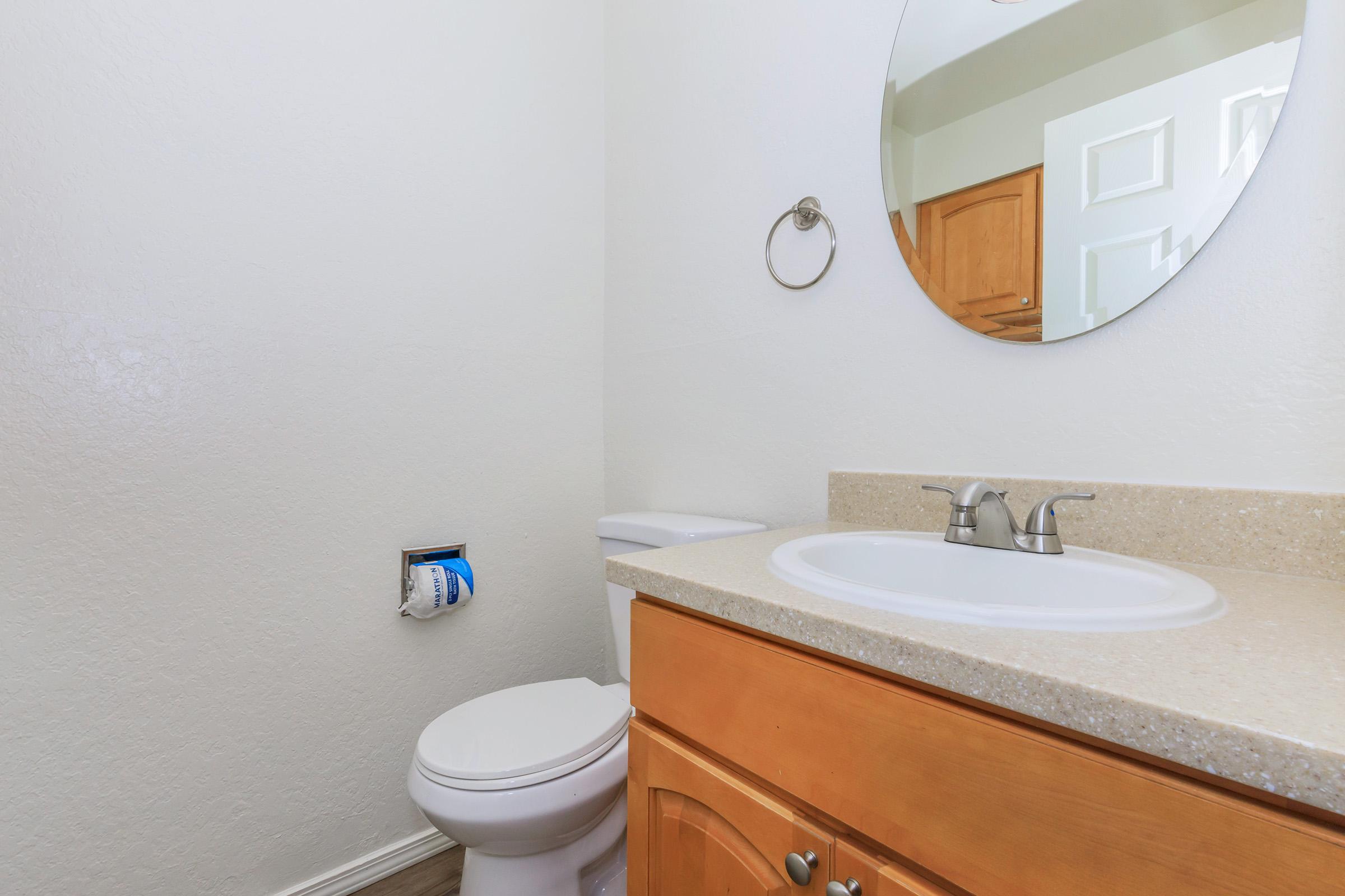 A clean, simple bathroom featuring a round mirror above a beige countertop with a sink. There is a toilet beside the sink, and a towel ring on the wall. The walls are painted white, and there are wooden cabinets in the background.