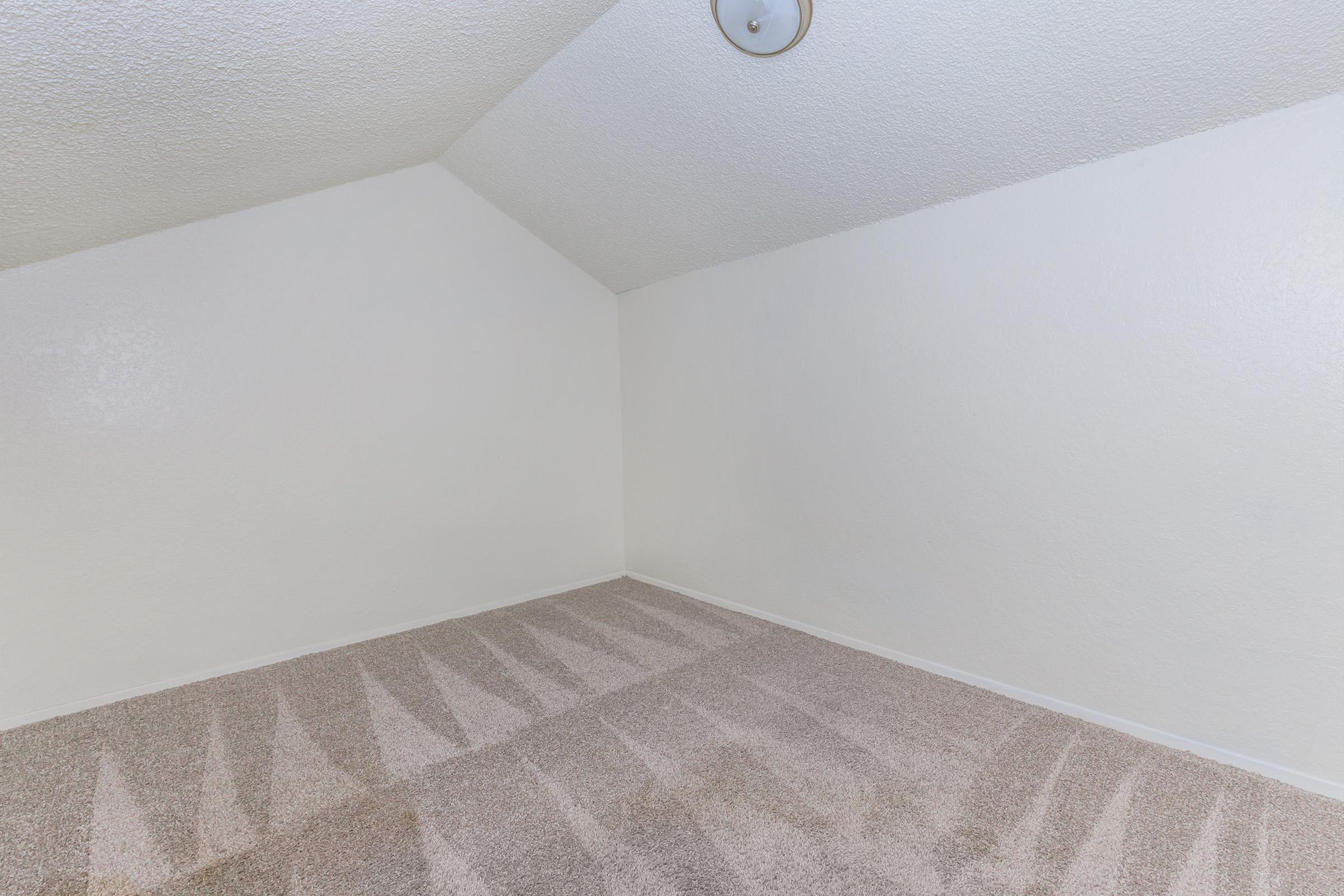 Empty room with sloped ceiling and neutral-colored walls. The carpet is light beige and shows subtle shadows. A simple ceiling light fixture is visible. The space appears bright and clean, suitable for various uses such as a bedroom or office.