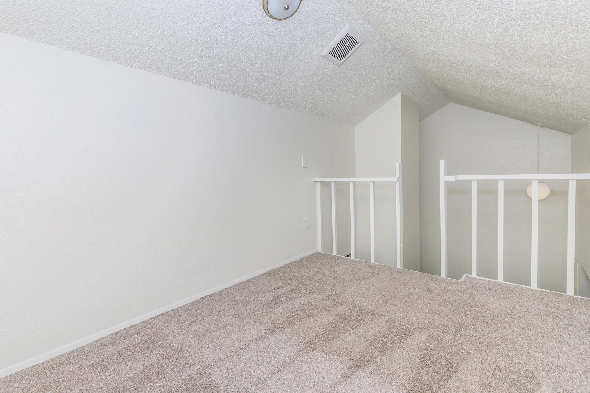 A well-lit, empty loft space with a sloped ceiling and beige carpet. There is a white railing along the edge, and natural light comes from a nearby window. The walls are painted white, giving the area a clean and spacious feel.