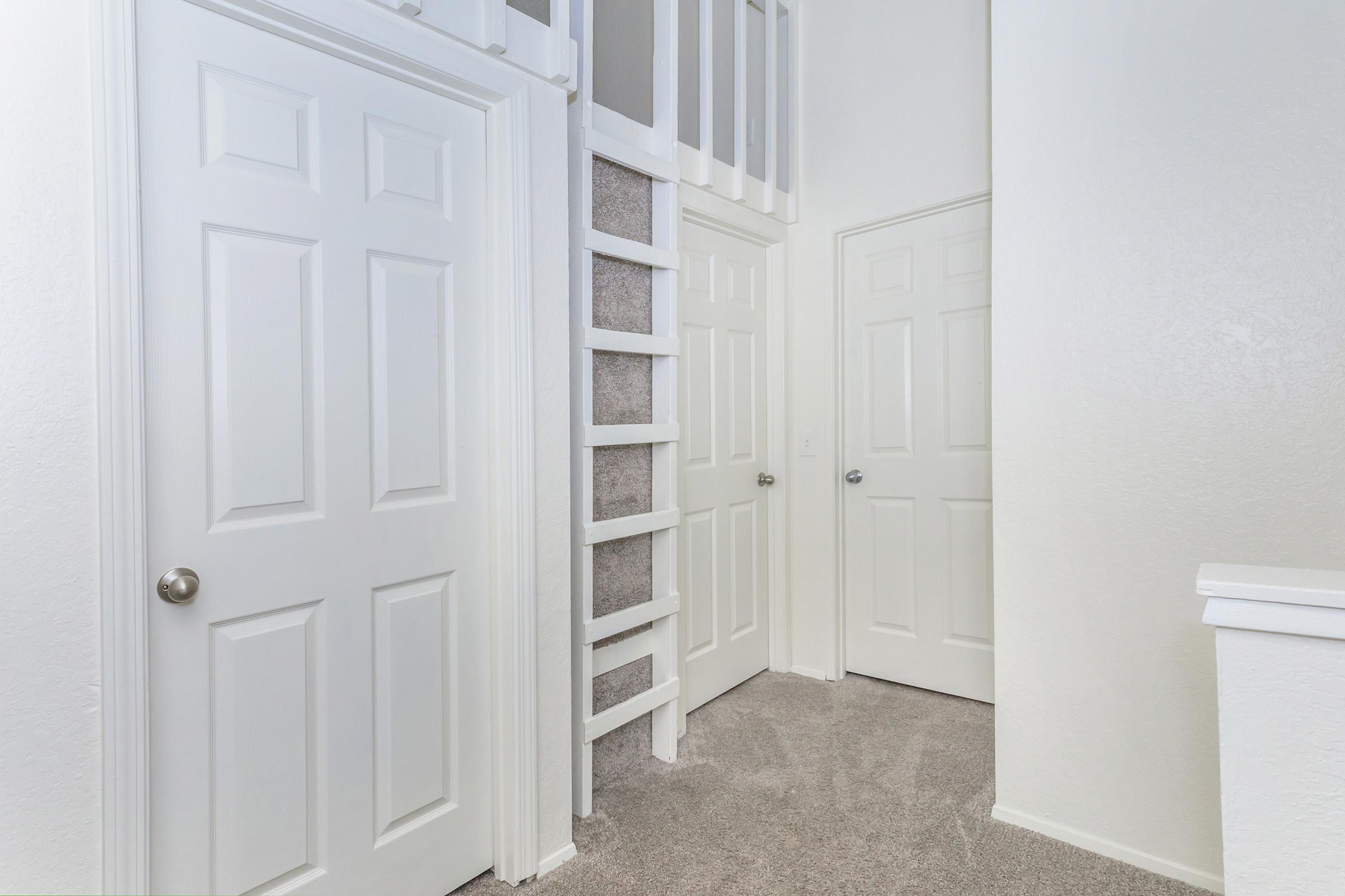 A well-lit hallway featuring three closed white doors, alongside a staircase leading to a loft area. The walls are painted in a light color, and the carpeted floor adds a cozy touch to the space.