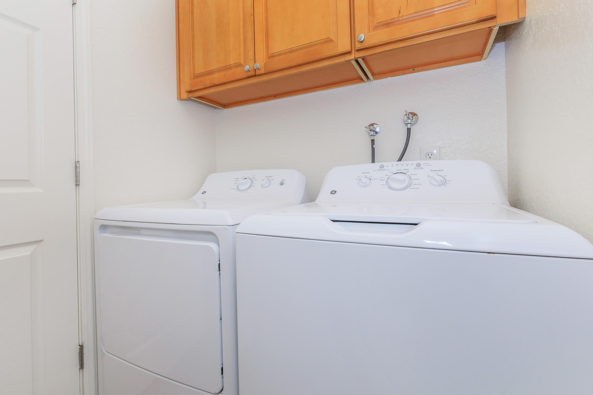A white washing machine and dryer side by side in a laundry room, with wooden cabinets above. The machines are positioned against a light-colored wall, and the room appears clean and organized.