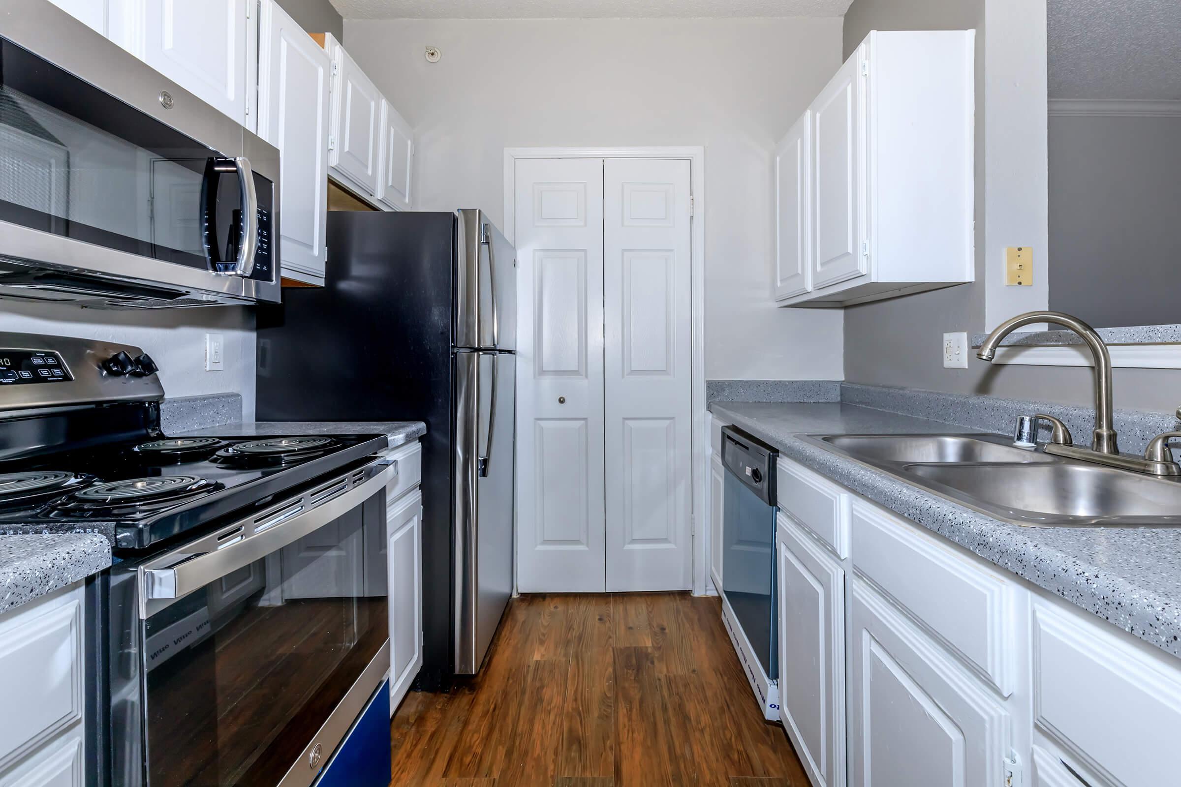 A modern kitchen featuring stainless steel appliances, including a microwave, oven, and refrigerator, alongside white cabinetry and a double sink. The countertops are gray, and the flooring is wood, creating a contemporary and inviting atmosphere. The room includes a closed door leading to another area.