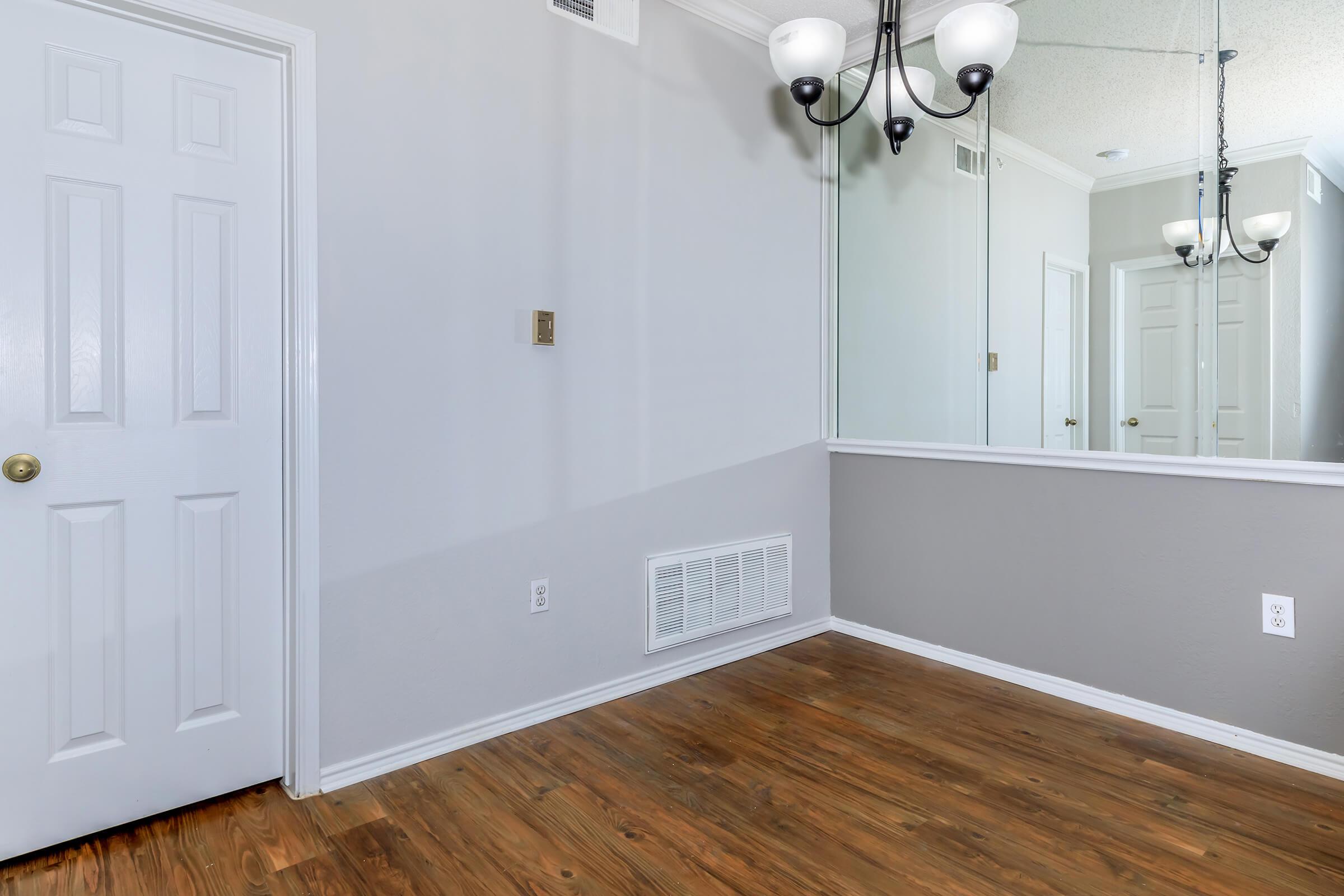 A well-lit room featuring wooden flooring and light gray walls. There's a mirror reflecting part of the room, and a chandelier hangs from the ceiling. A closet door is visible on the left, and a vent is located by the wall. The space appears clean and modern, suggesting an inviting atmosphere.