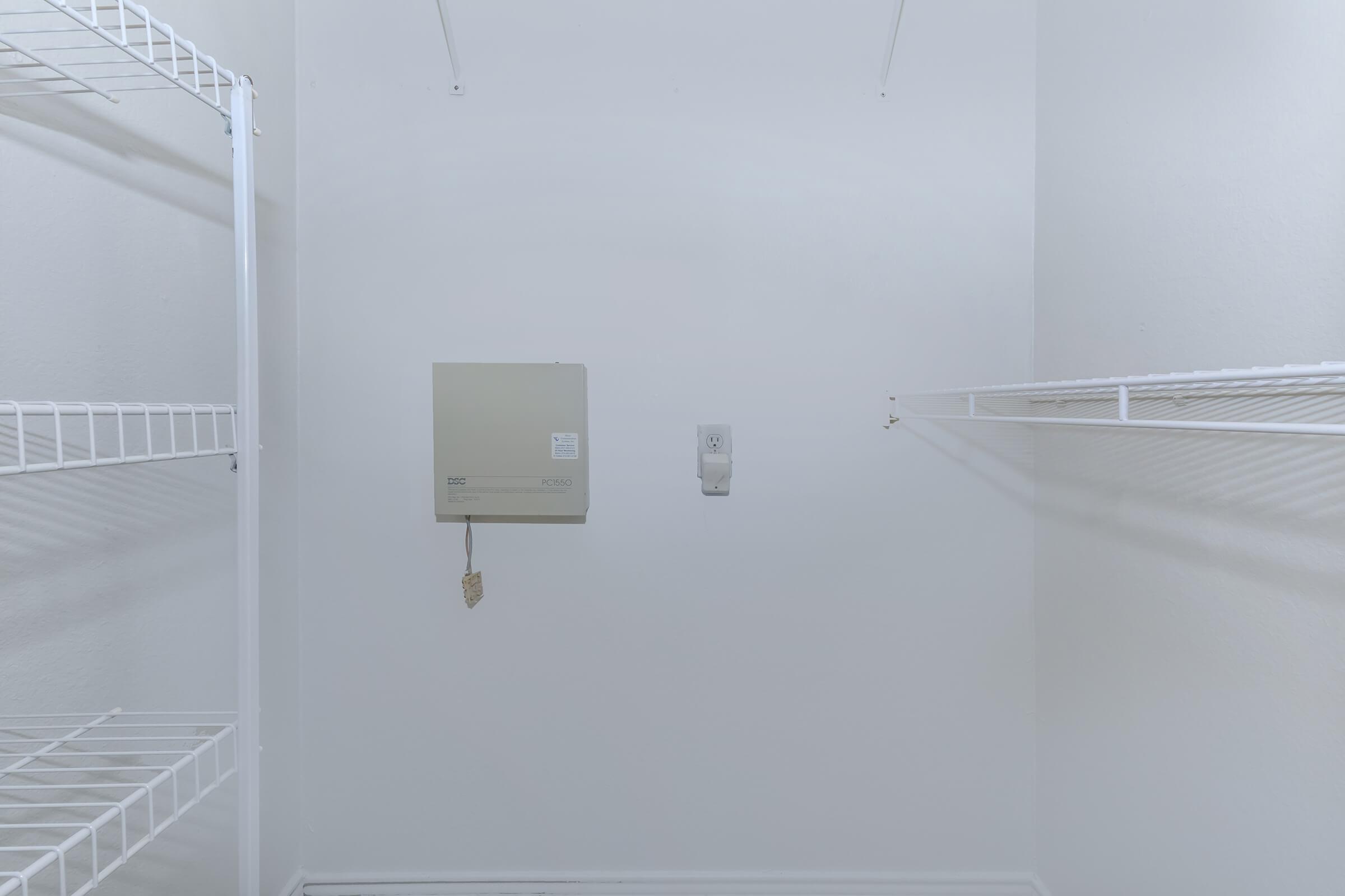 A minimalist empty closet featuring white wire shelving on both sides. A beige electrical panel is mounted on the left wall, with a small white device positioned nearby. The walls and floor are plain white, contributing to a clean, uncluttered aesthetic.