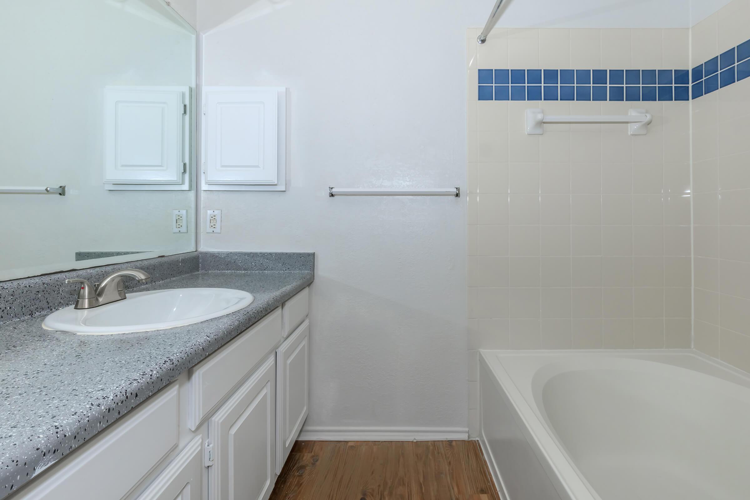 A clean and modern bathroom featuring a countertop sink with a mirror above, white cabinetry, and a bathtub. The wall tiles are beige with a blue accent strip. Wooden flooring complements the design, and a towel bar is mounted on the wall, creating a bright and spacious atmosphere.