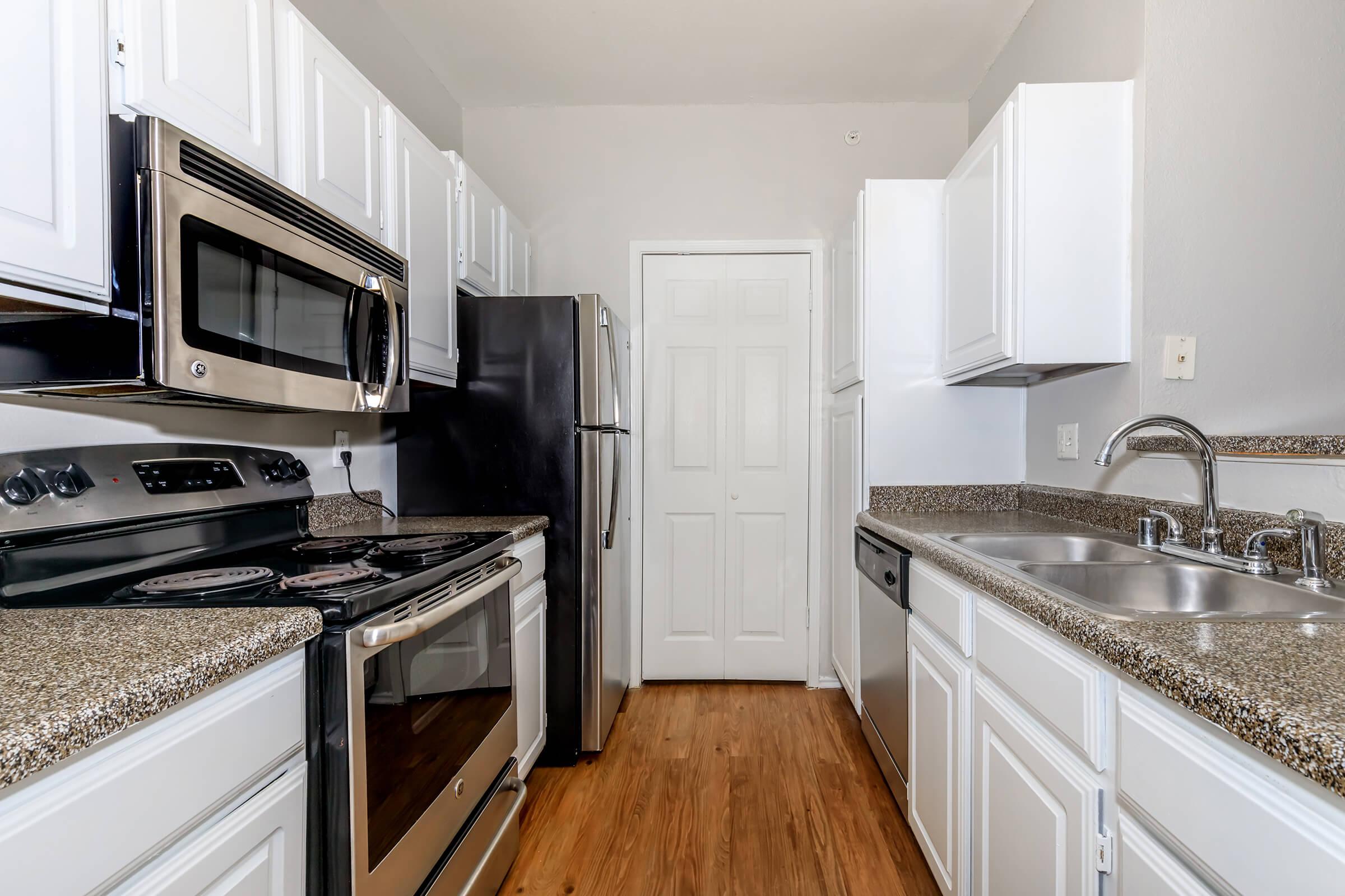 A modern kitchen featuring stainless steel appliances, including a microwave, oven, and refrigerator. The cabinets are white, and the countertops are a light-colored speckled material. The floor is wooden, and there are dual sinks with a faucet. The area is well-lit and organized, creating a welcoming cooking space.