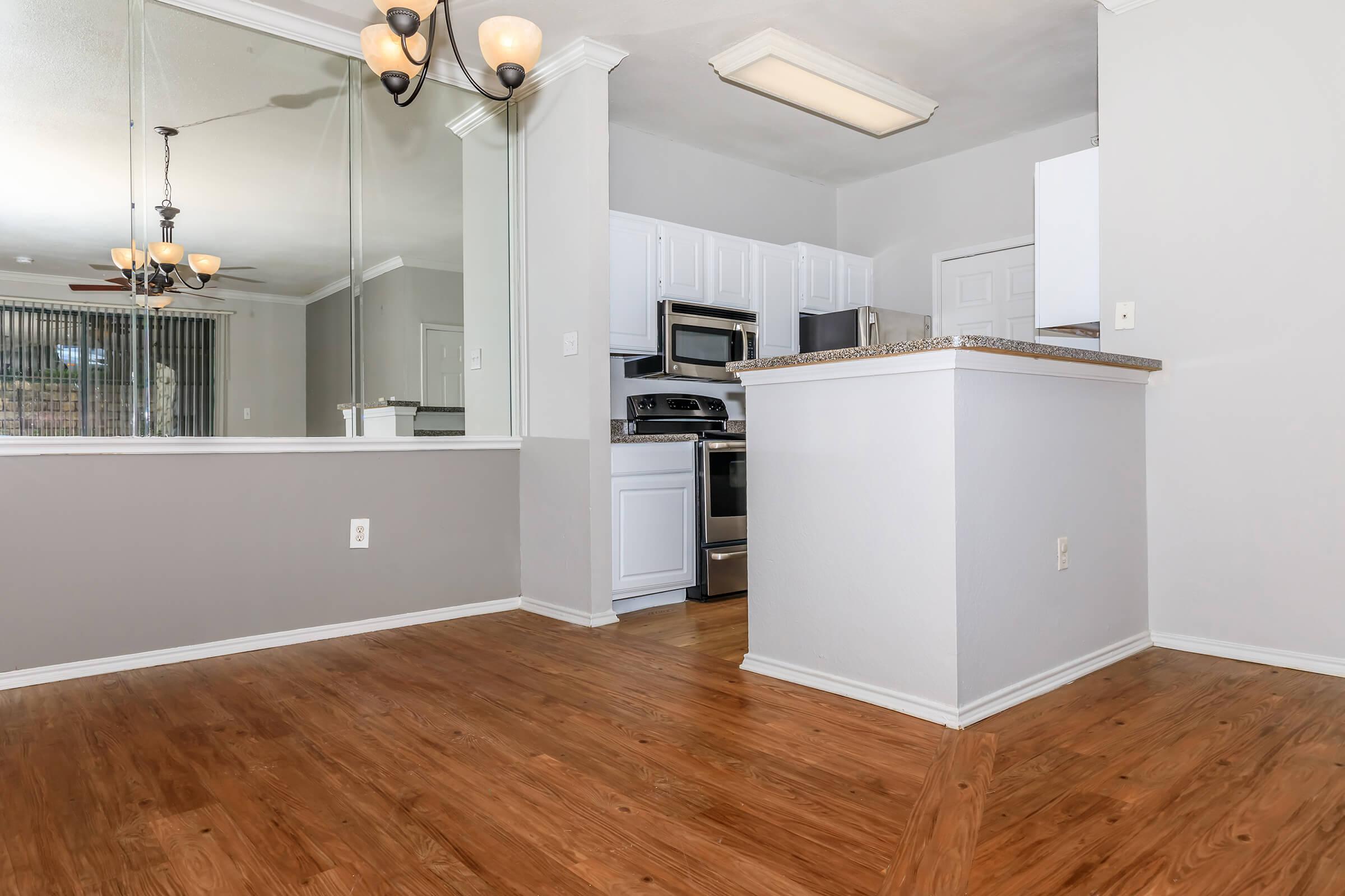 View of a modern kitchen and dining area featuring a light-colored wall, hardwood flooring, and mirrored accents. The kitchen includes stainless steel appliances, white cabinetry, and a countertop that separates it from the dining space. A chandelier hangs from the ceiling, adding a warm ambiance.