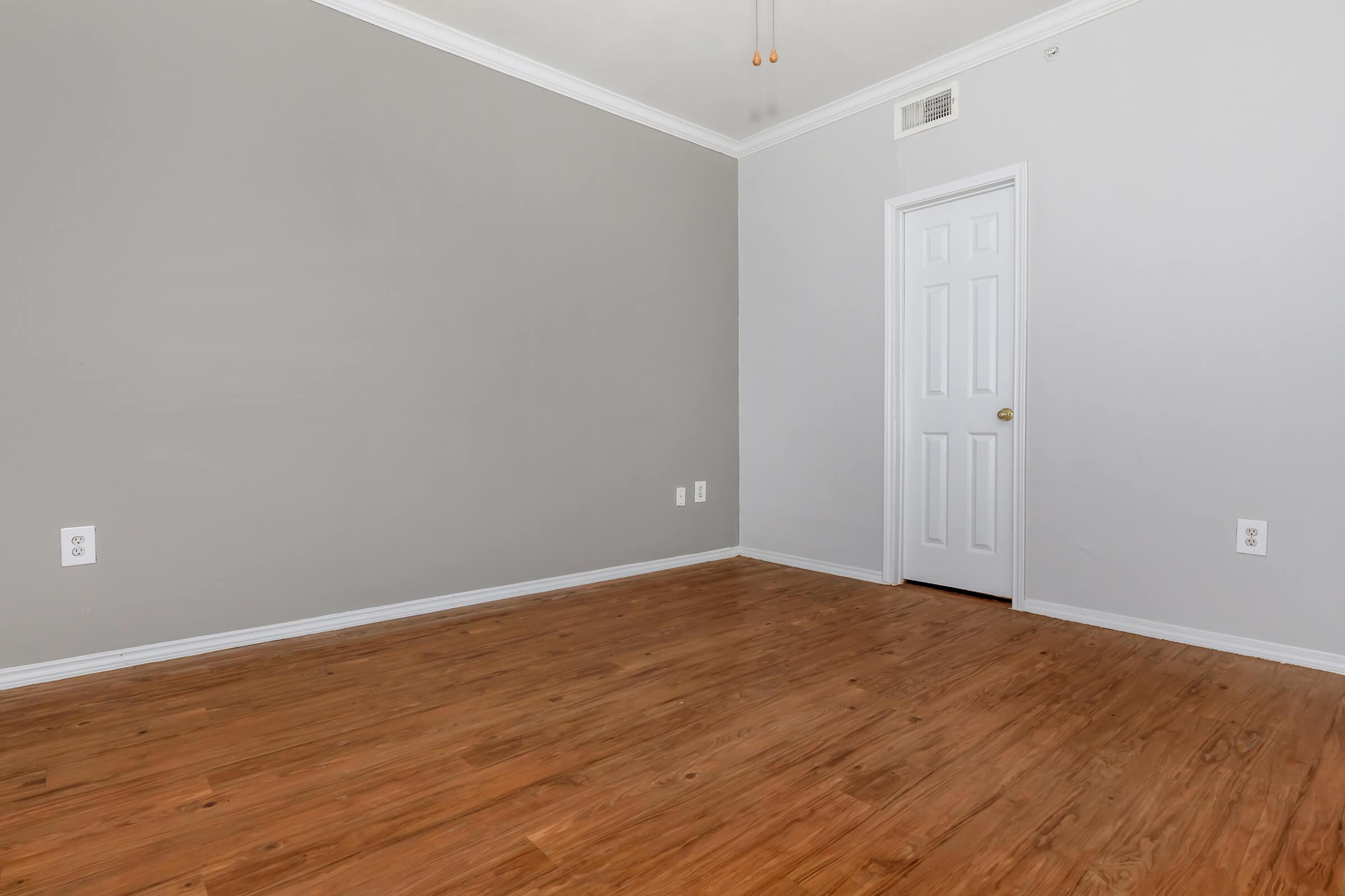 A minimalistic room with neutral gray walls and wooden flooring. One side features a closed white door and a light switch on the wall. The space is empty and evokes a clean, simple aesthetic with natural light possibly illuminating the interior.