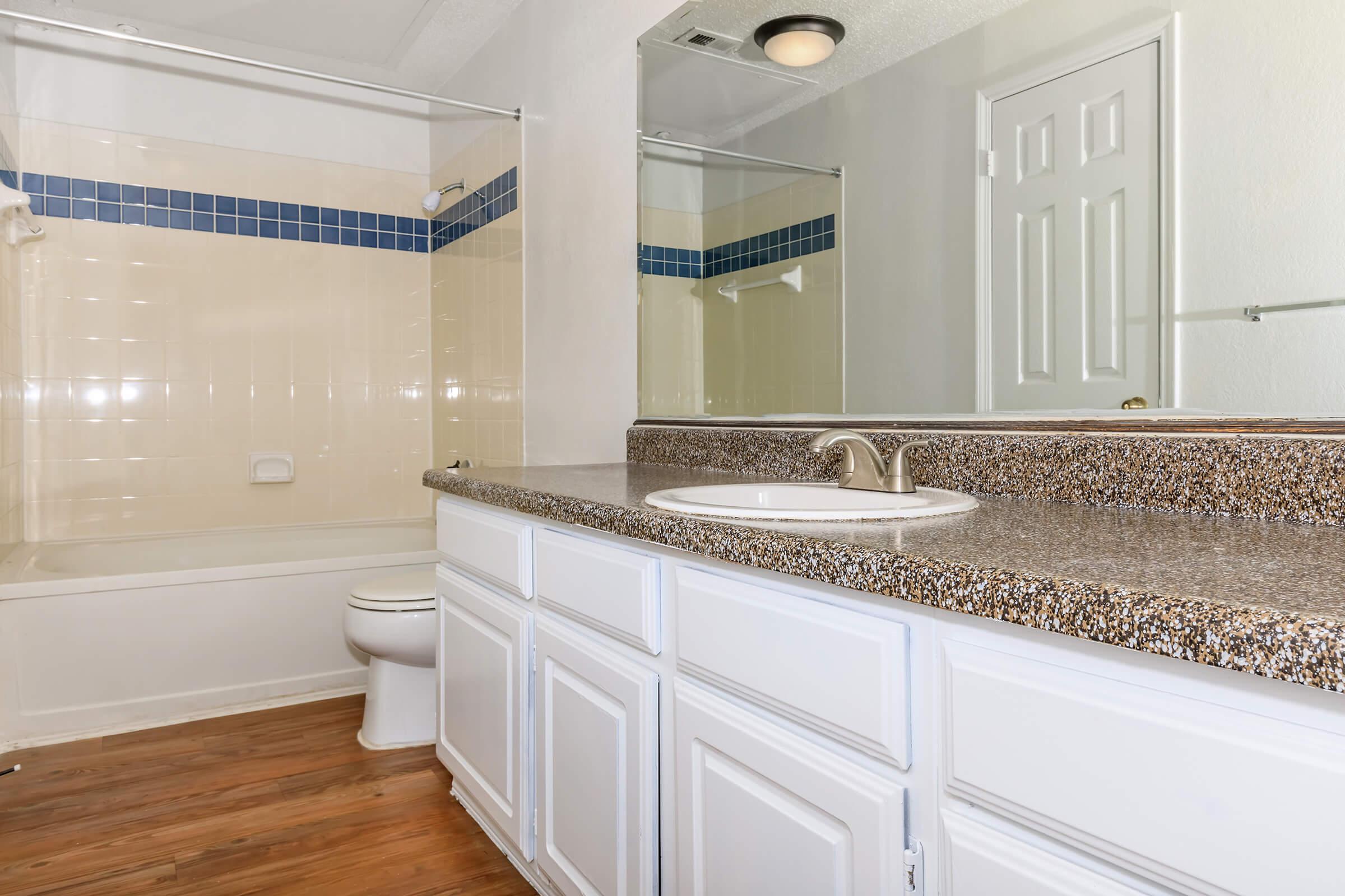 Modern bathroom featuring a spacious vanity with a sink, a large wall mirror, and a shower/tub combo. The walls are tiled with a light cream color and accented with blue tiles. The floor is wooden, adding warmth to the space. Clean and well-lit environment, ideal for a contemporary home.