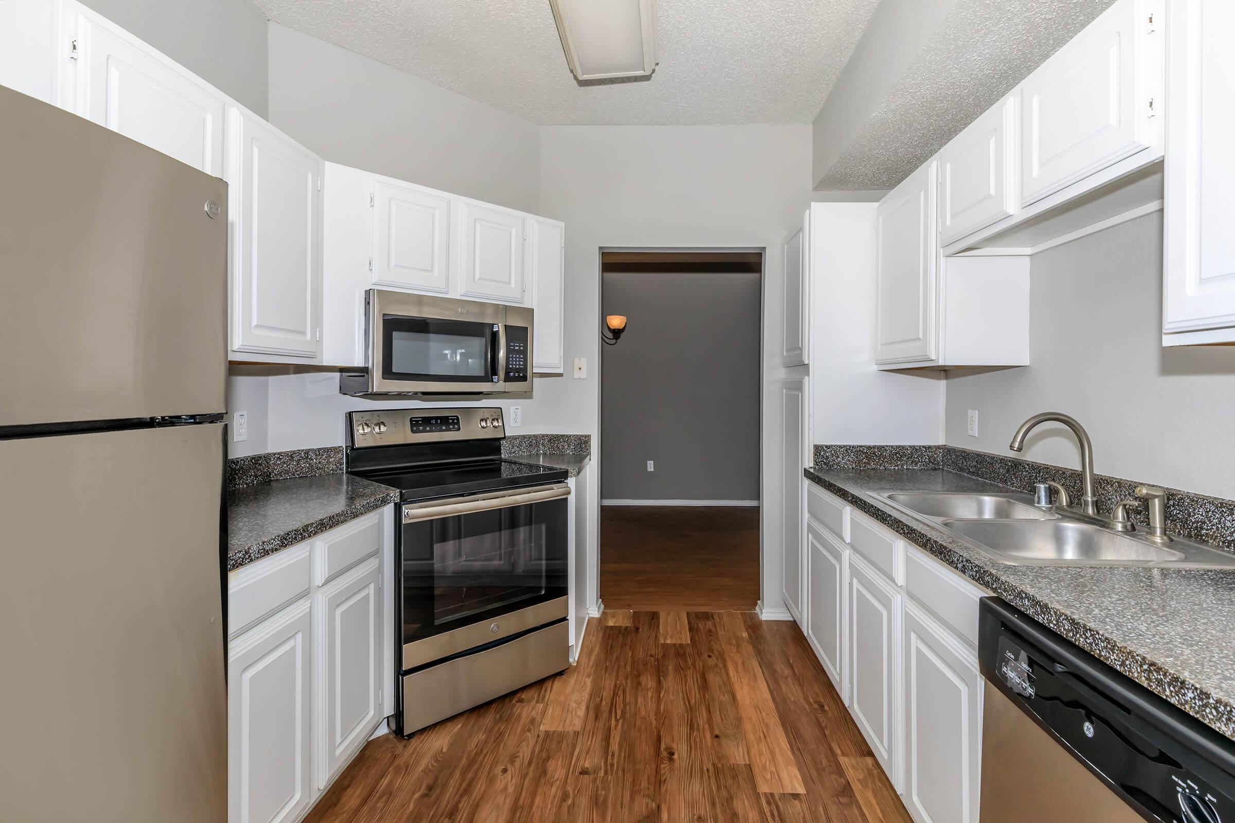 A modern kitchen featuring stainless steel appliances, including a refrigerator, microwave, and stove, surrounded by white cabinetry and a granite countertop. The flooring is wood-like laminate. The room is well-lit with a neutral color scheme, leading to a doorway in the background.