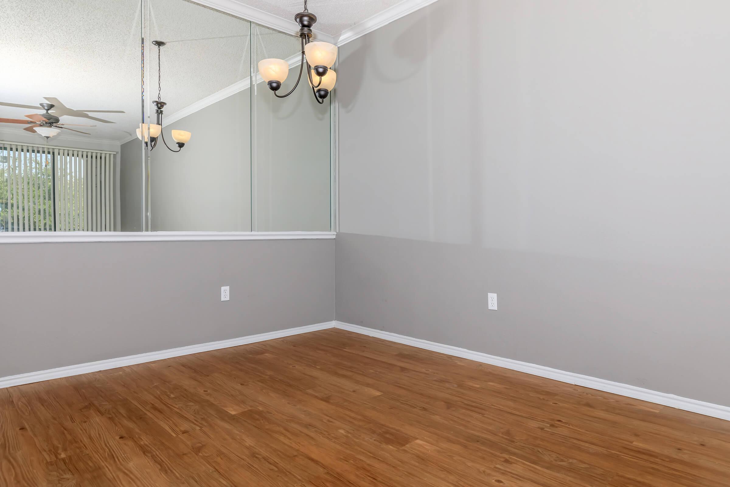 A spacious room with light gray walls featuring a mirror on one side. The floor has warm-toned wood, and there are three hanging light fixtures with a vintage design. A ceiling fan is visible in the background, creating a bright and airy ambiance. Natural light filters in through nearby windows.