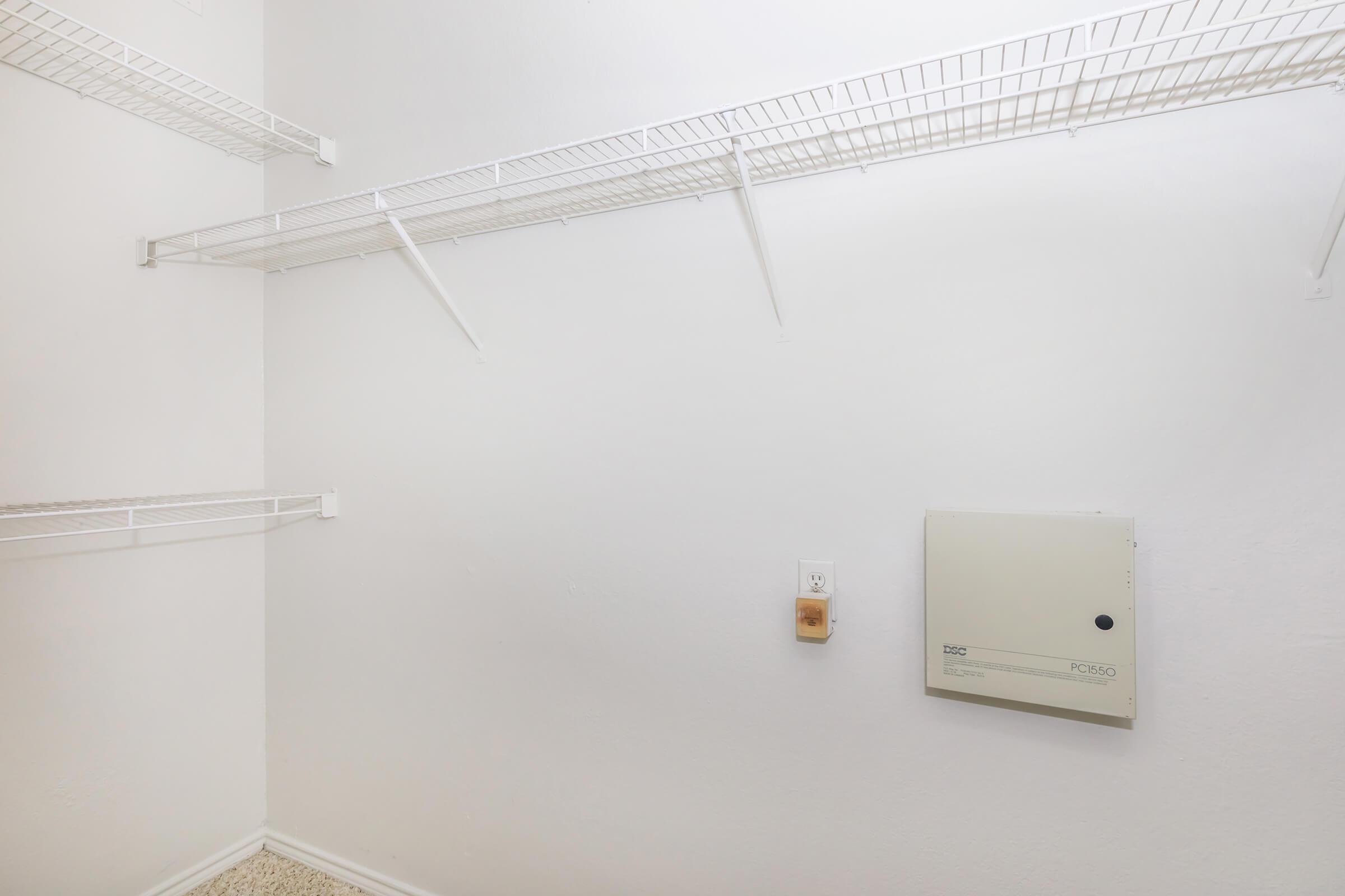 A clean, empty closet with white walls and two wire shelves on either side. The floor is carpeted, and there is an electrical panel mounted on the wall. The space is well-lit and organized, ideal for storage.