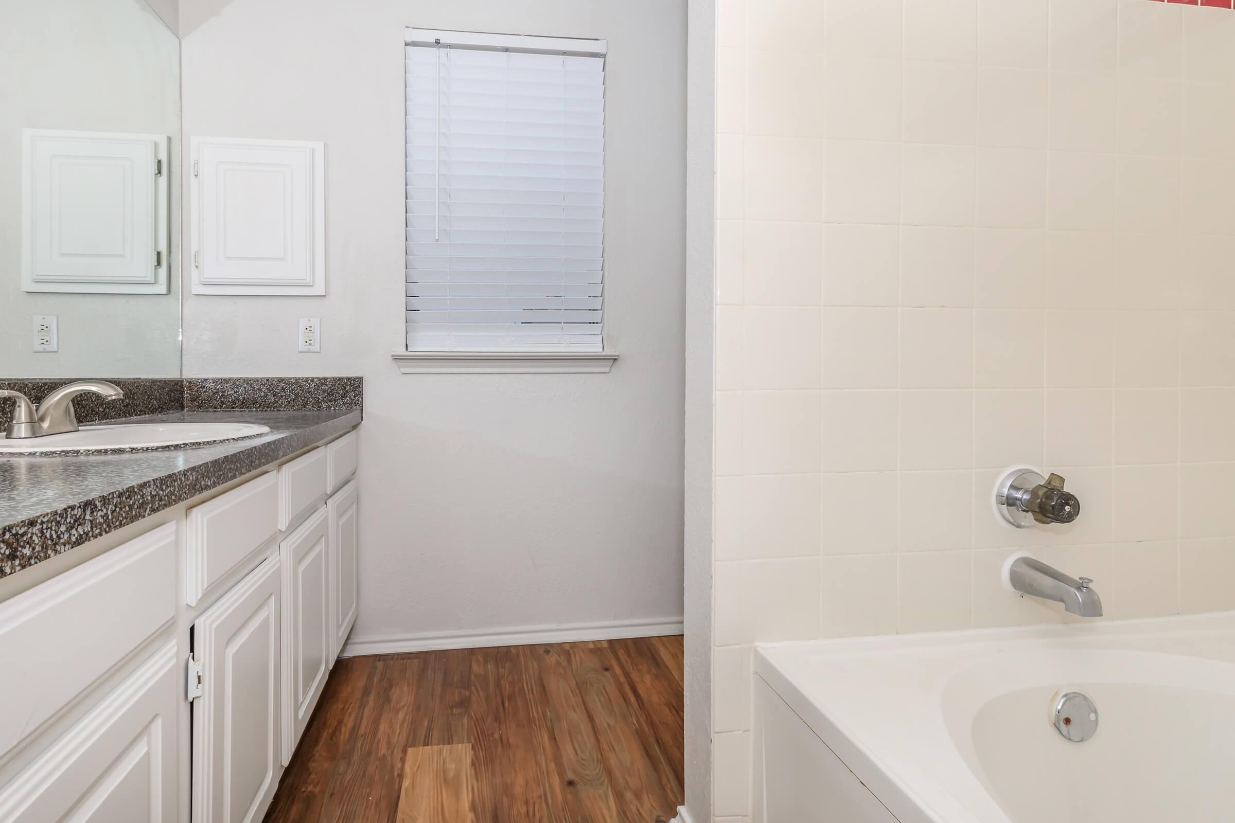 A well-lit bathroom featuring a white bathtub, a granite countertop with dual sinks, and a large mirror. The floor is made of wood, and there is a window with blinds providing natural light. The walls are painted in a light color, creating an airy and spacious feel.