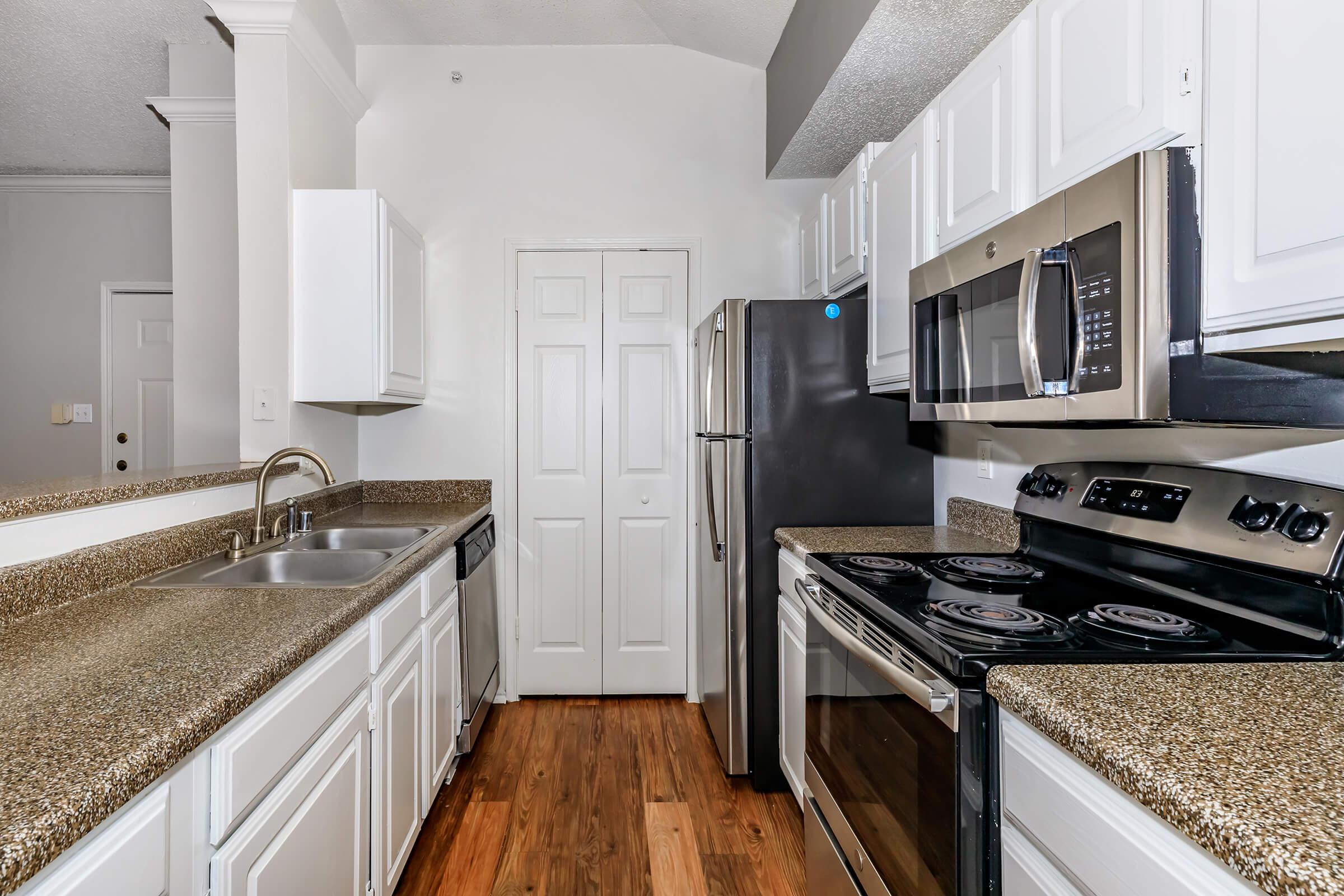 A modern kitchen featuring white cabinetry, granite countertops, stainless steel appliances, including an oven, microwave, and refrigerator. The kitchen has a double sink with a faucet, and the flooring is made of wood. The walls are painted white, creating a bright and airy atmosphere.