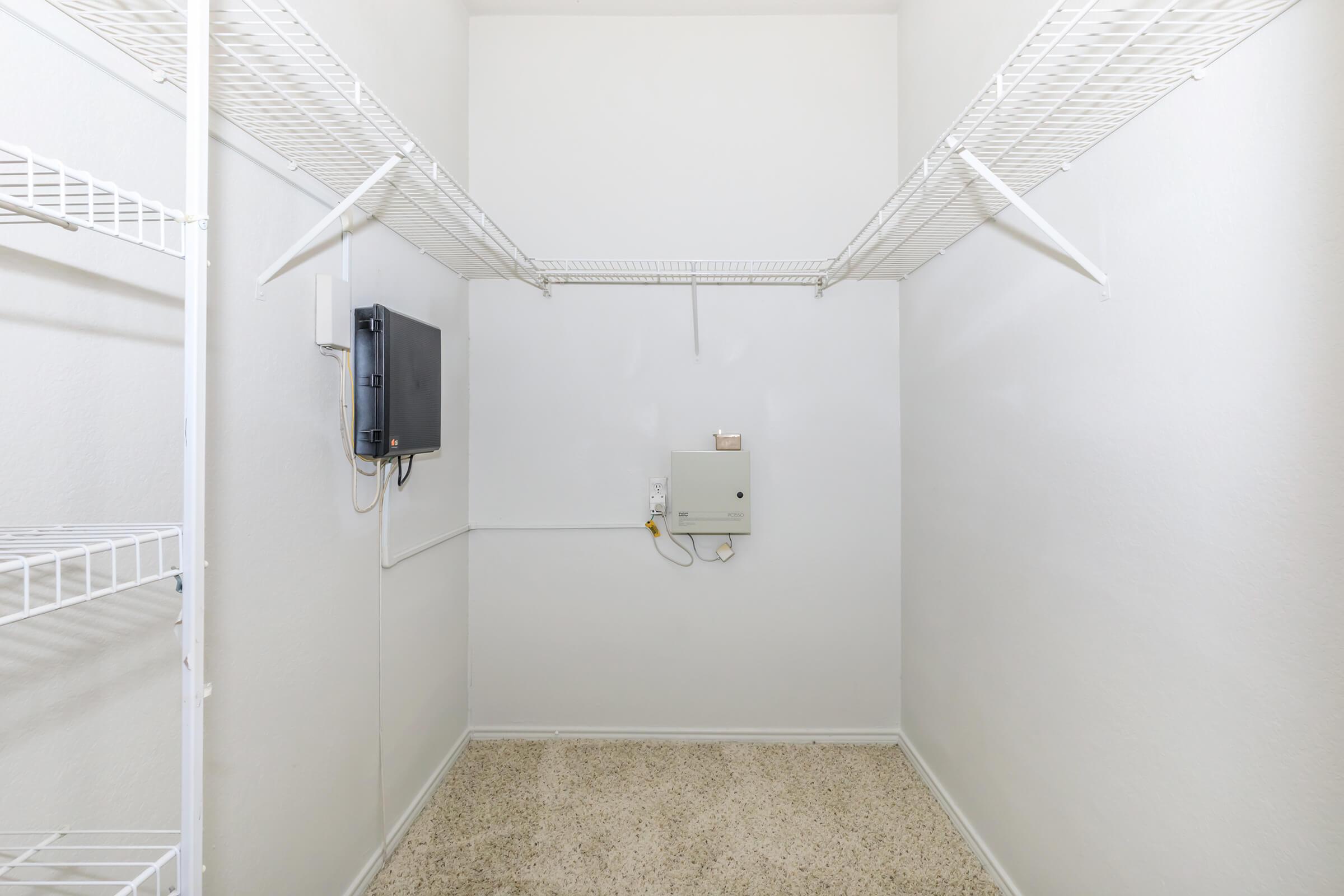 Empty utility room featuring white wire shelving on the left and right walls. There is a control panel mounted on the back wall and a small device near it. The walls and floor are painted in neutral tones, creating a clean, well-lit space.