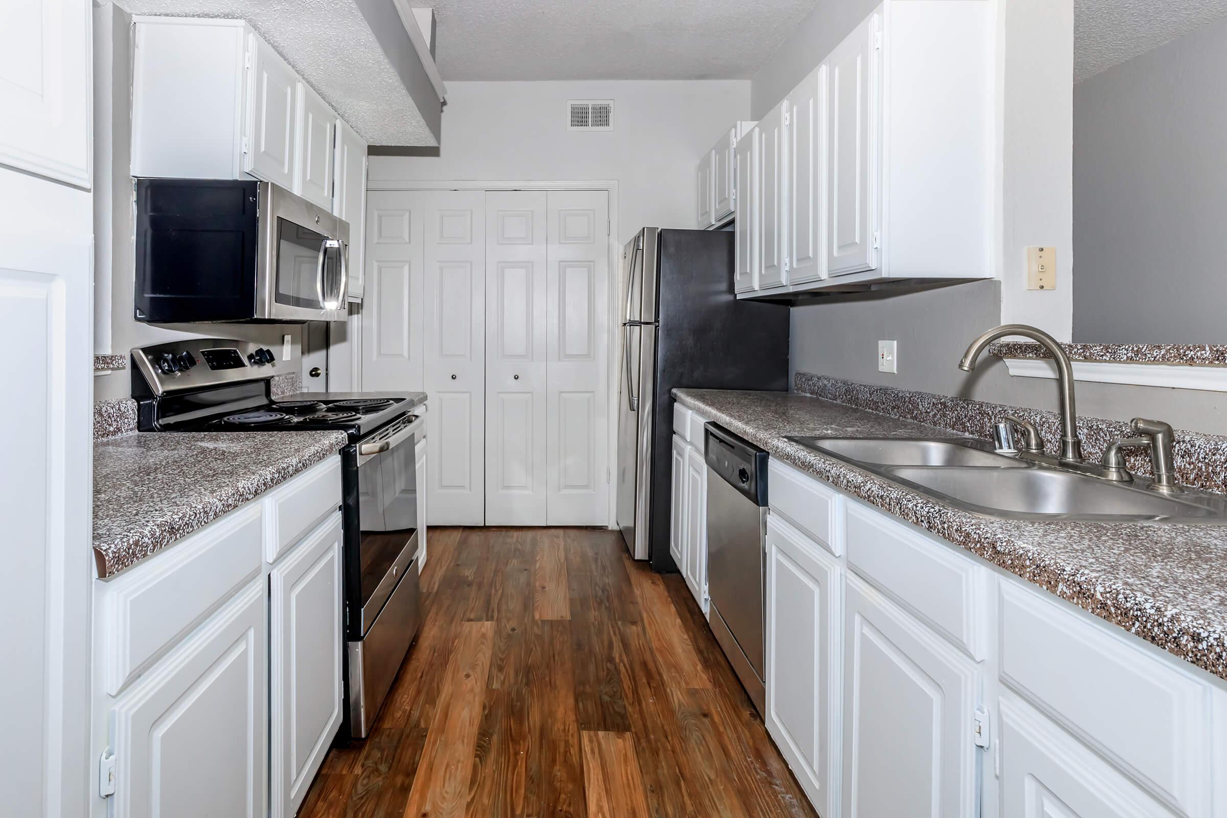 A modern kitchen featuring white cabinetry, stainless steel appliances, and granite countertops. The kitchen includes a gas stove, microwave, dishwasher, and a double sink, with a wooden floor and neutral-colored walls. Two closed doors are visible at the back, suggesting storage or a pantry.