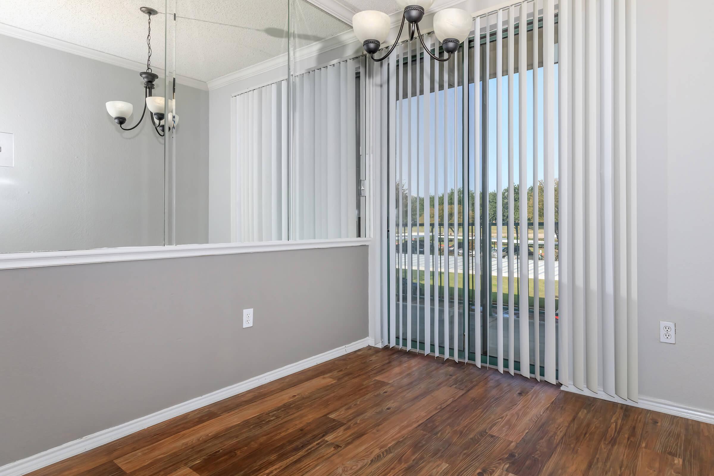 A well-lit interior space featuring a wooden floor and light gray walls. A large window with vertical blinds allows natural light to enter. A decorative chandelier hangs from the ceiling, and there is a mirrored wall on one side, enhancing the sense of openness in the room.