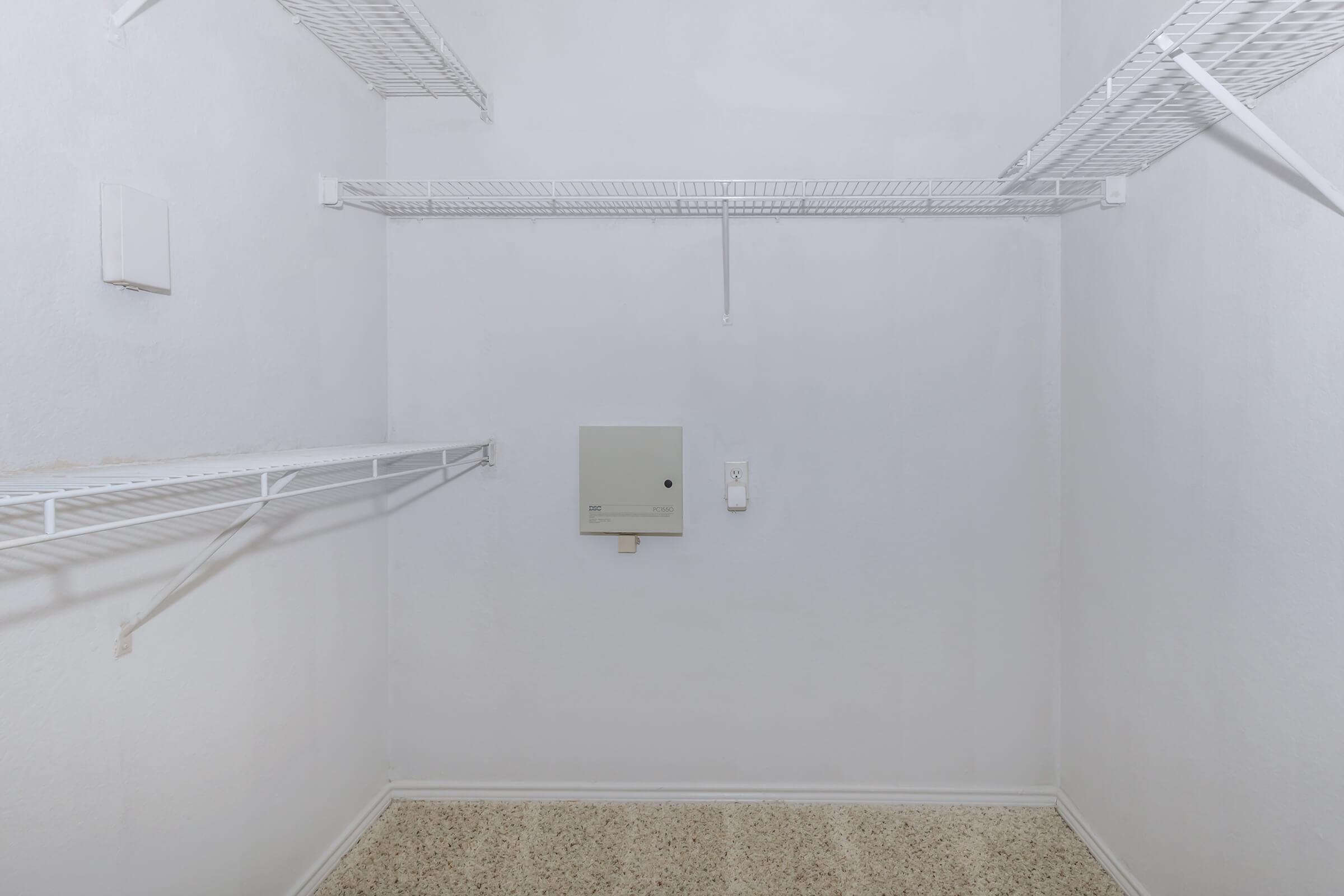 Empty closet with white wire shelving on two walls. The walls and floor are painted in light colors. A small electrical box is mounted on one wall, and there is a light switch nearby. The overall space is clean and uncluttered.