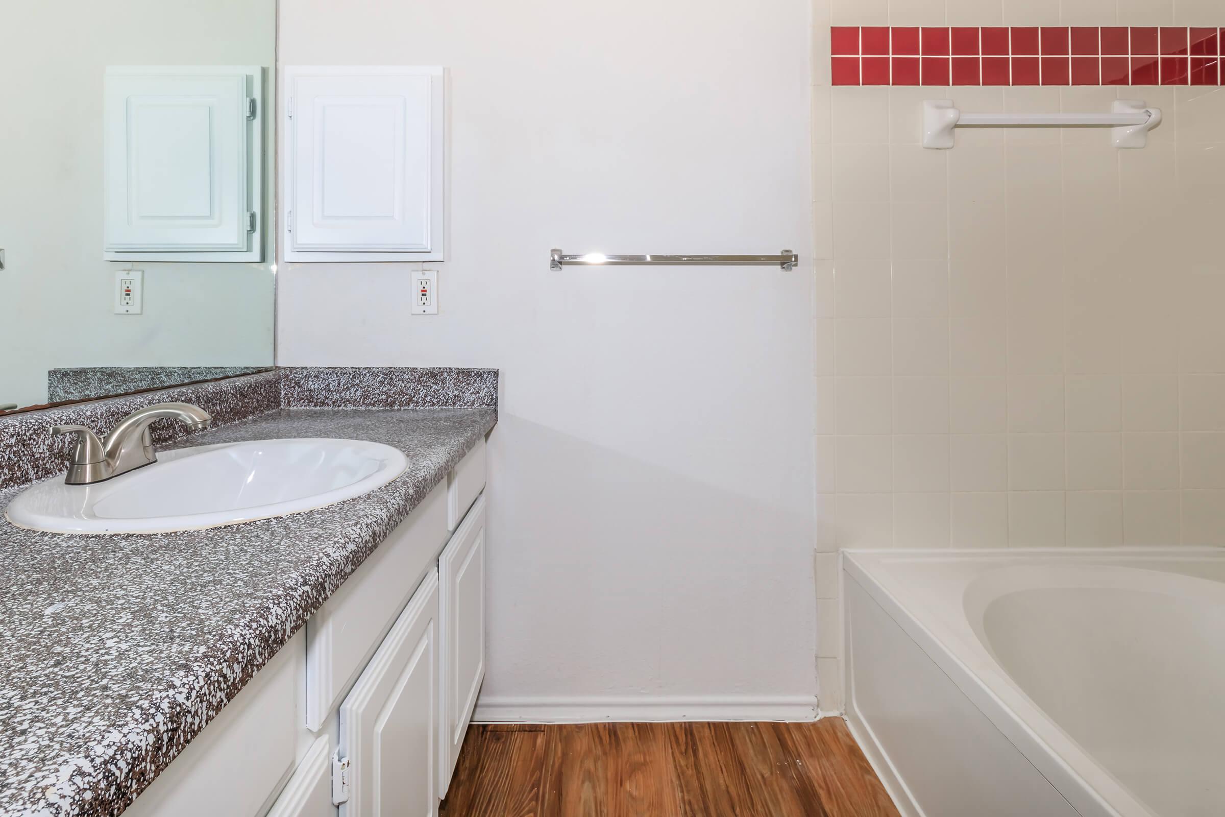 A clean, modern bathroom featuring a sink with a granite countertop, a large mirror, a towel rack, and a bathtub. The wall has red tiles along the top, and there are light wood floors. The overall color scheme is neutral, creating a simple and inviting atmosphere.