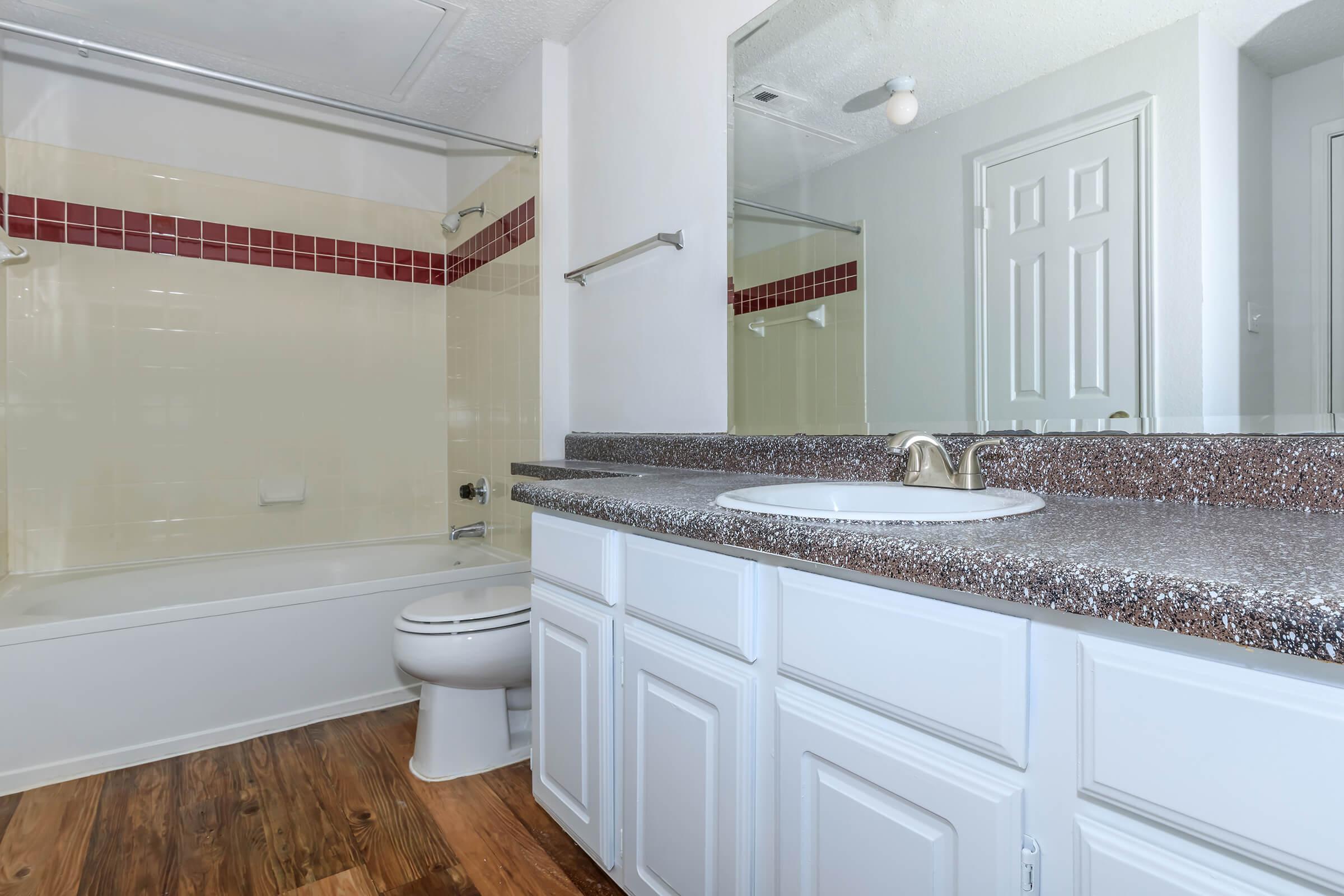 A clean and modern bathroom featuring a white bathtub and shower combination, a spacious countertop with a sink, and a white cabinetry. The walls are light-colored, with a decorative tile border in shades of red. Wooden flooring adds warmth to the space, and there is a mirror above the sink.