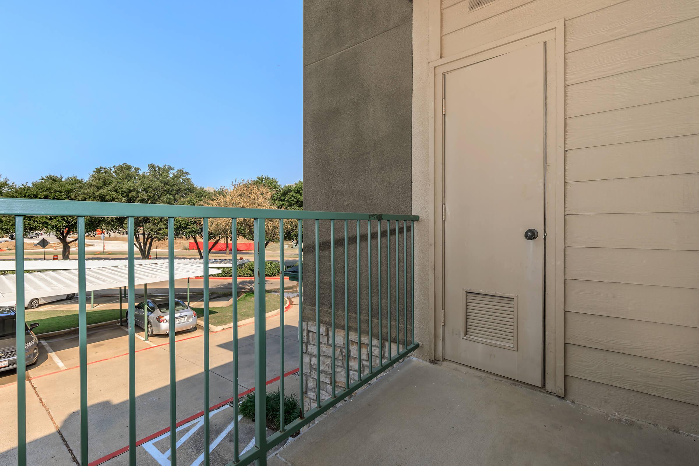 A clear view from a balcony overlooking a parking lot, featuring a green metal railing and a closed door on the neighboring wall. The scene is bright and sunny, with trees visible in the background and parked cars below.