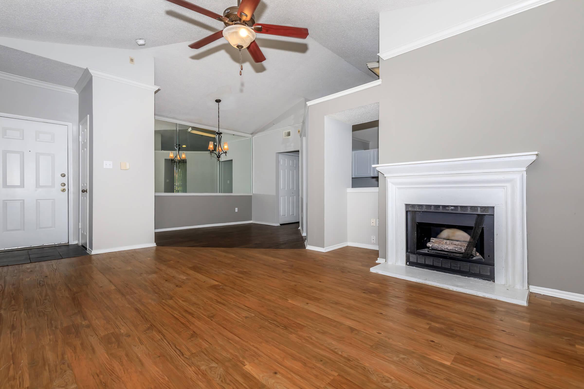 A spacious living room with a vaulted ceiling and ceiling fan, featuring a modern fireplace with a white surround. Light gray walls and hardwood floors create a warm atmosphere. To the left, there's a front door, while a dining area is visible in the background with a glass wall separating it from the kitchen.