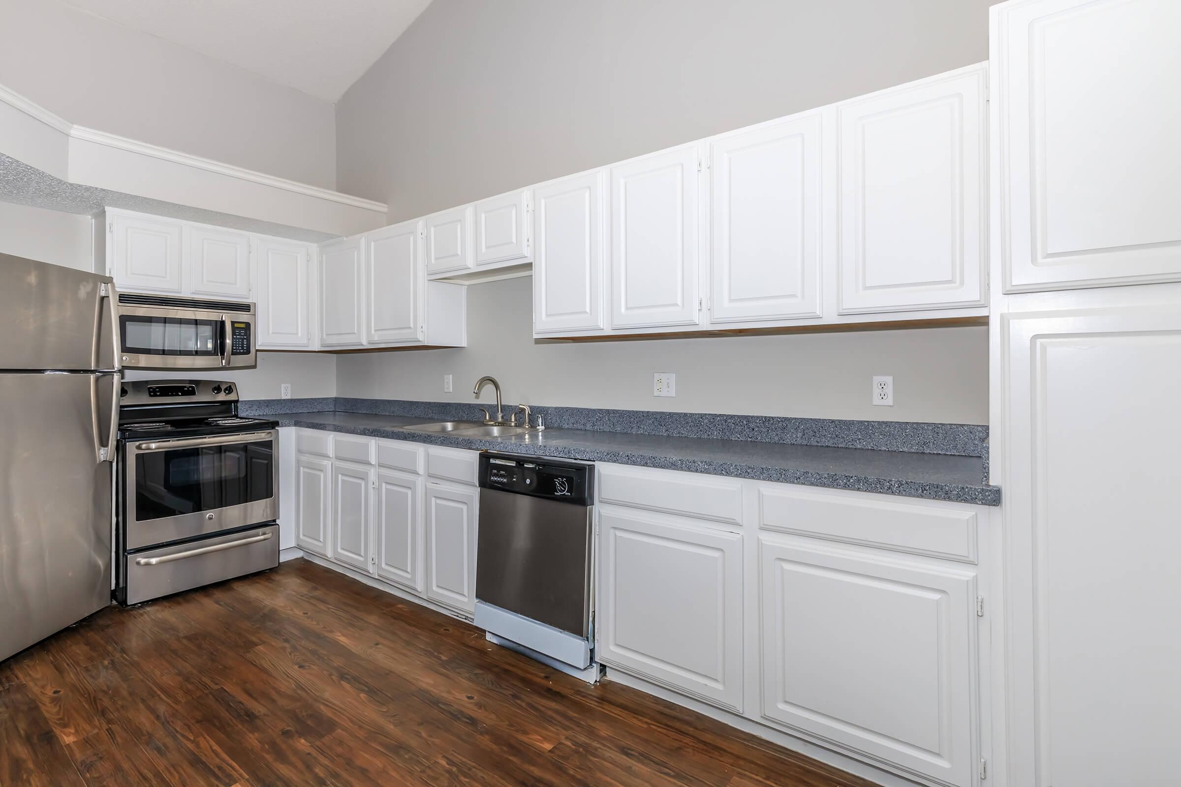 Modern kitchen with white cabinetry, stainless steel appliances including a refrigerator, oven, and dishwasher. The countertops are grey, and the flooring is a warm, wooden tone. Bright walls and high ceilings contribute to a spacious, airy feel.