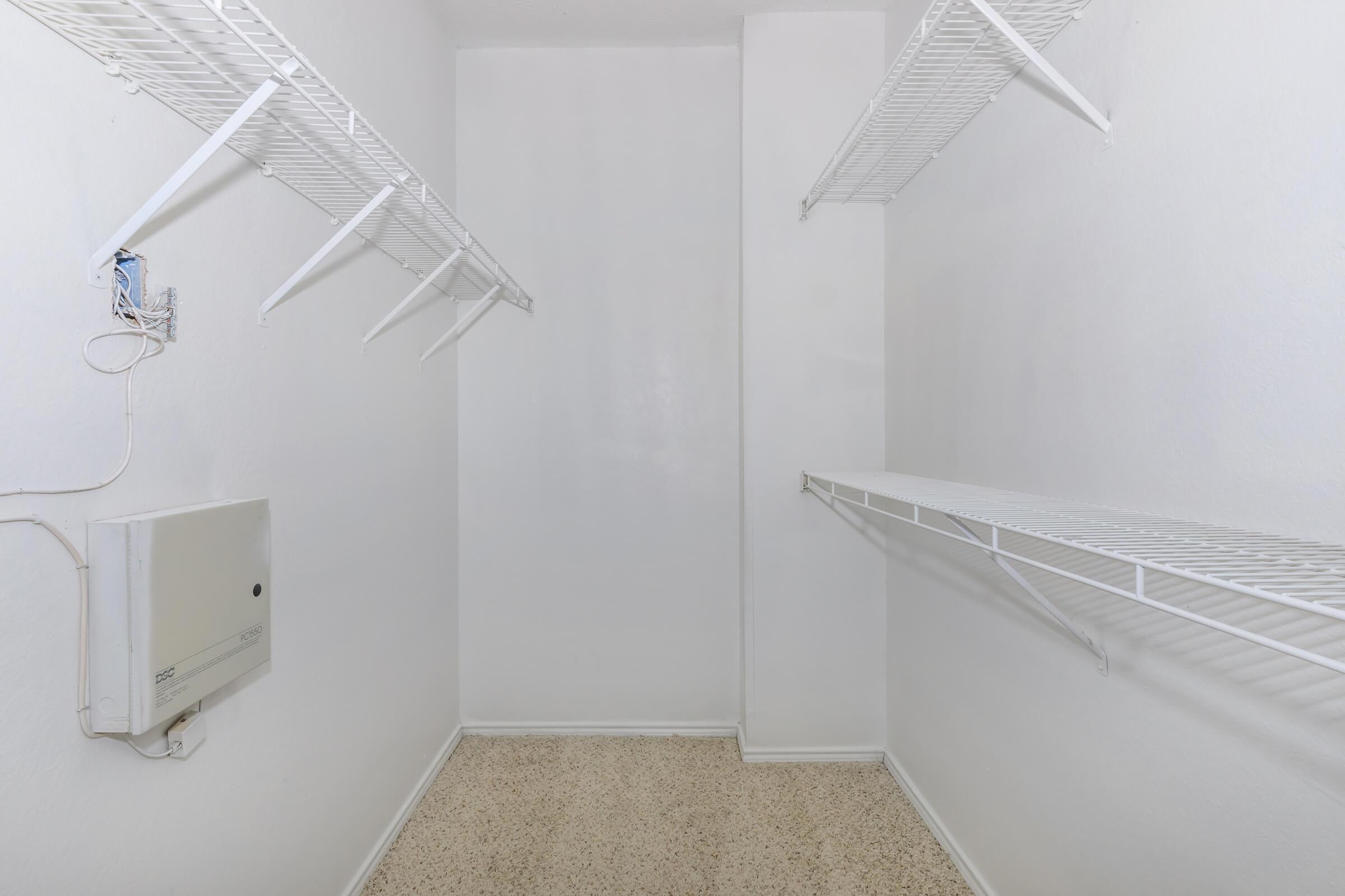 A clean, empty closet featuring white wire shelving on the walls. The walls are painted white, and the floor is light-colored with speckled texture. An electrical panel is mounted on one wall, and there are no items or decorations present in the space.