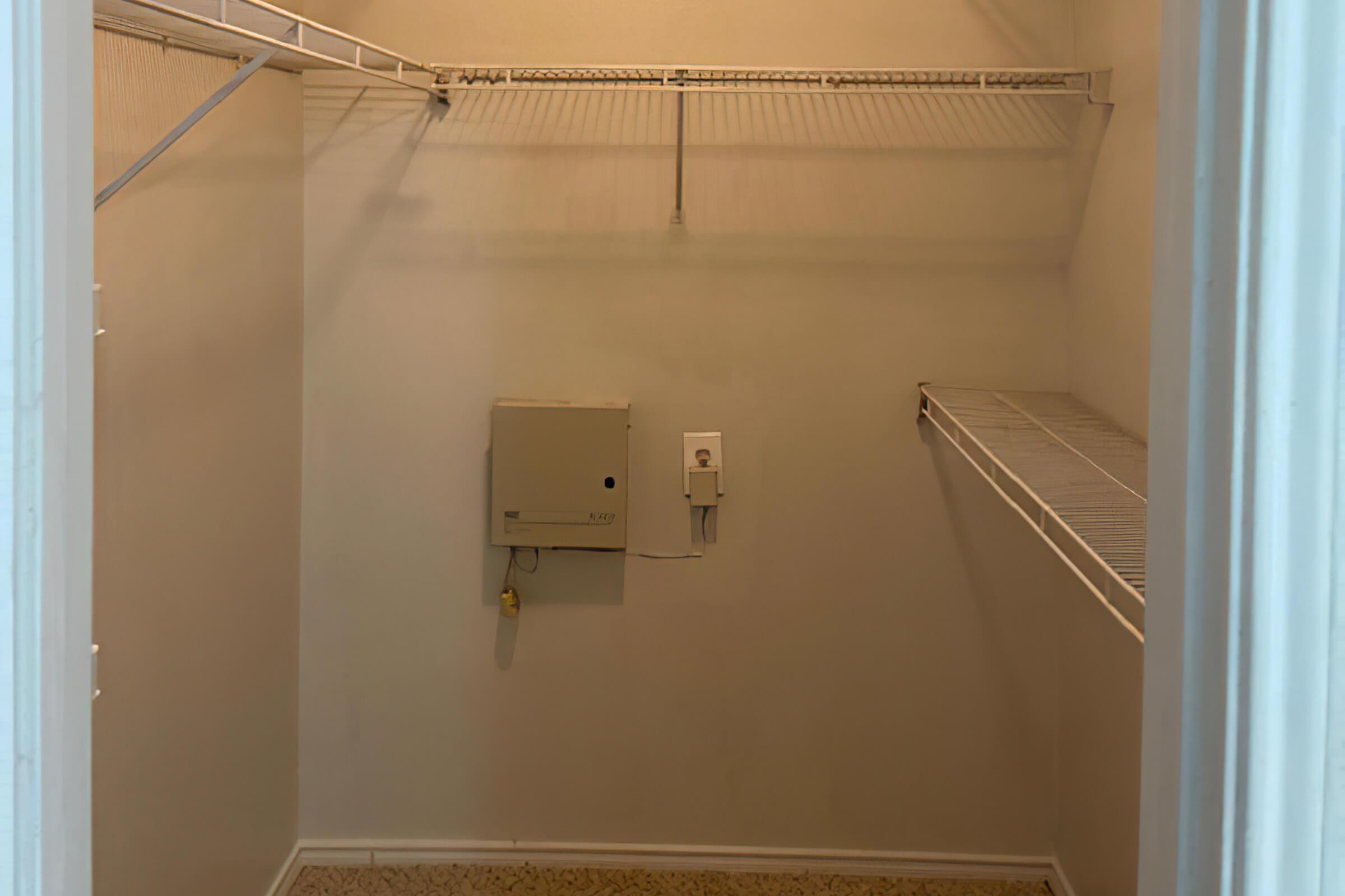 A small, empty closet with beige walls and a shelf running along the top. There is a white wire shelf on one side and an electrical panel mounted on the wall. The floor is carpeted in a neutral tone. The lighting is soft and there are no visible items or decorations in the space.